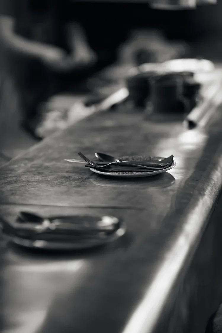 A black and white photo of a restaurant table with stacked plates, spoons, a knife, and bowls, with a blurred background.