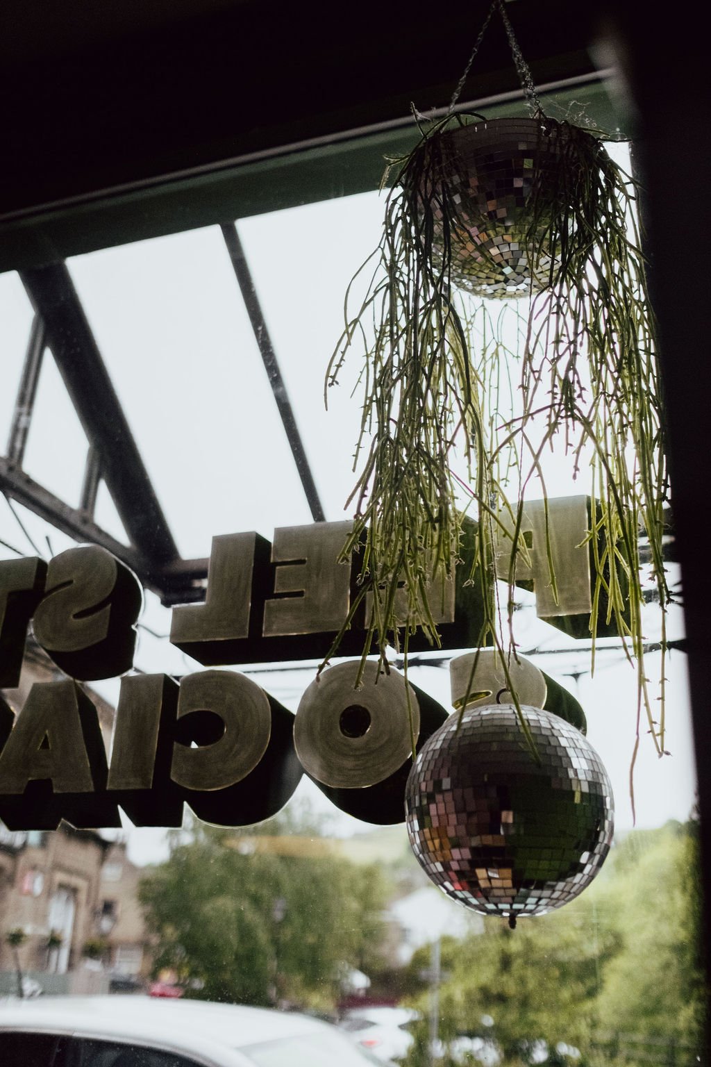 Hanging plants and disco balls are seen through a window with a sign that spells 'Welcome.' Outside, trees and parked cars are visible.