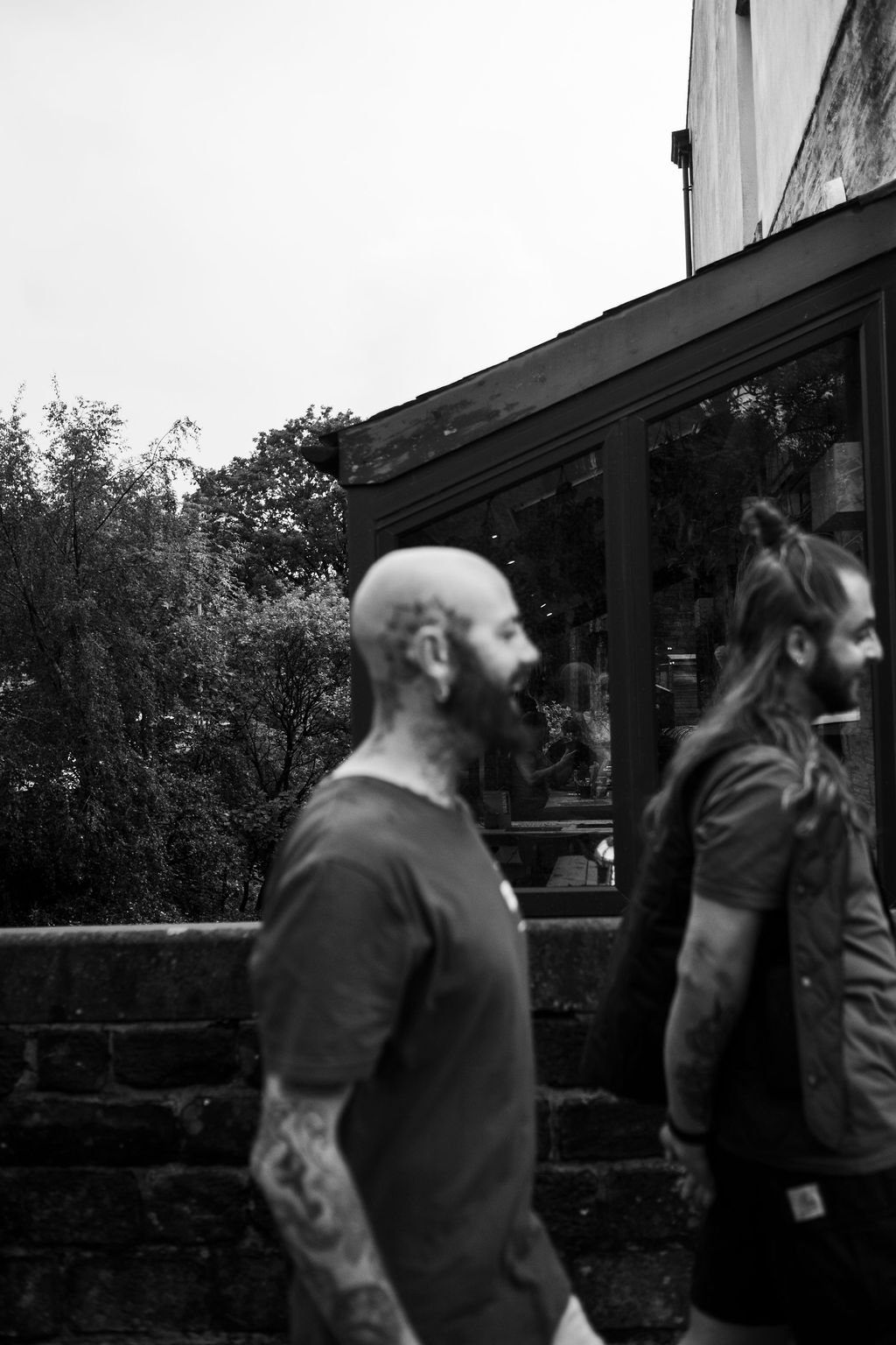 Two men walking side by side outdoors, one with a beard and tattoos, and the other with long hair tied up, in front of a large window with trees and a building reflected on it.