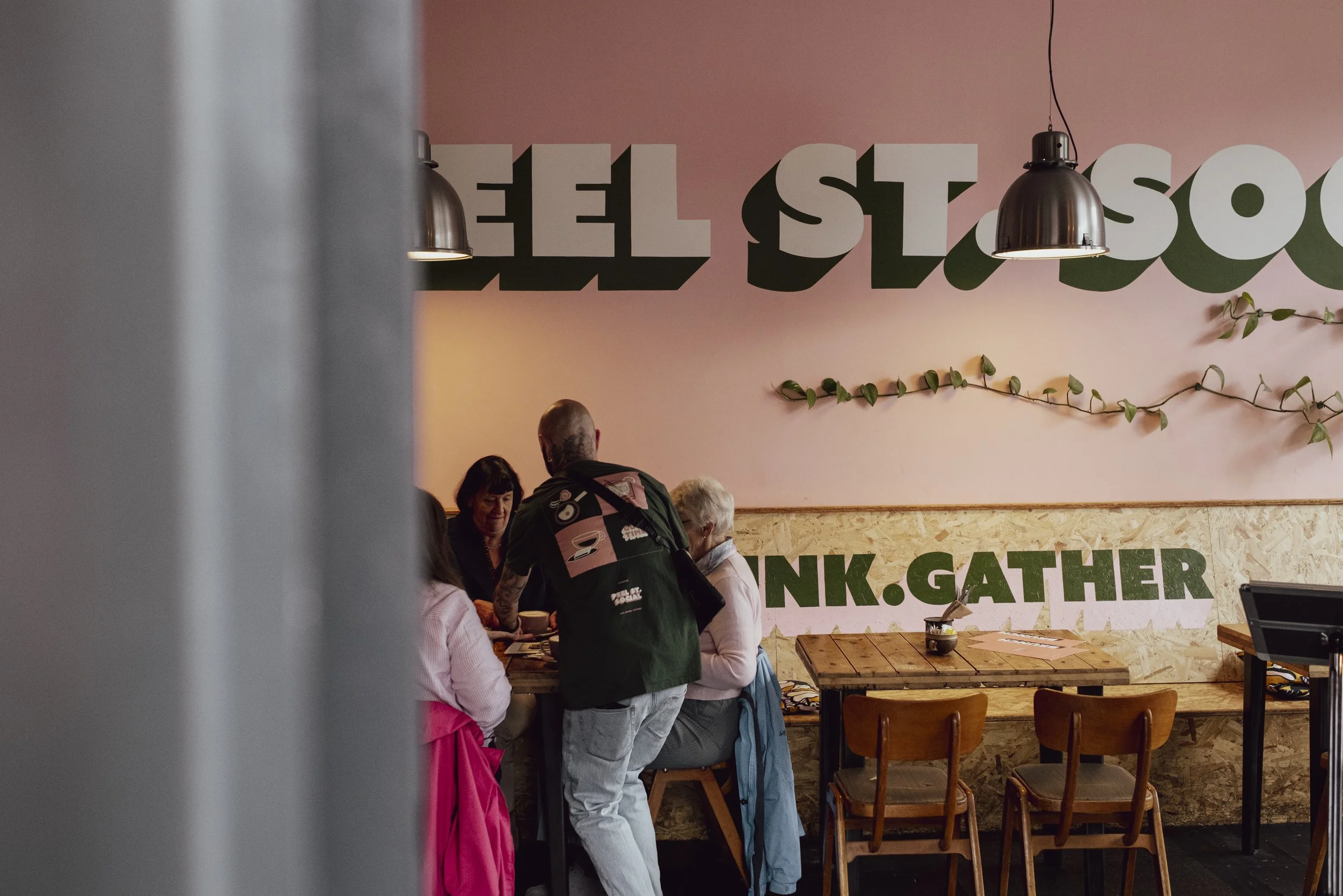 People gathered at a counter inside a restaurant with a pink wall and large white letters spelling 'ELLE' behind them. There are hanging lamps, a leafy vine decoration, and wooden tables and chairs.
