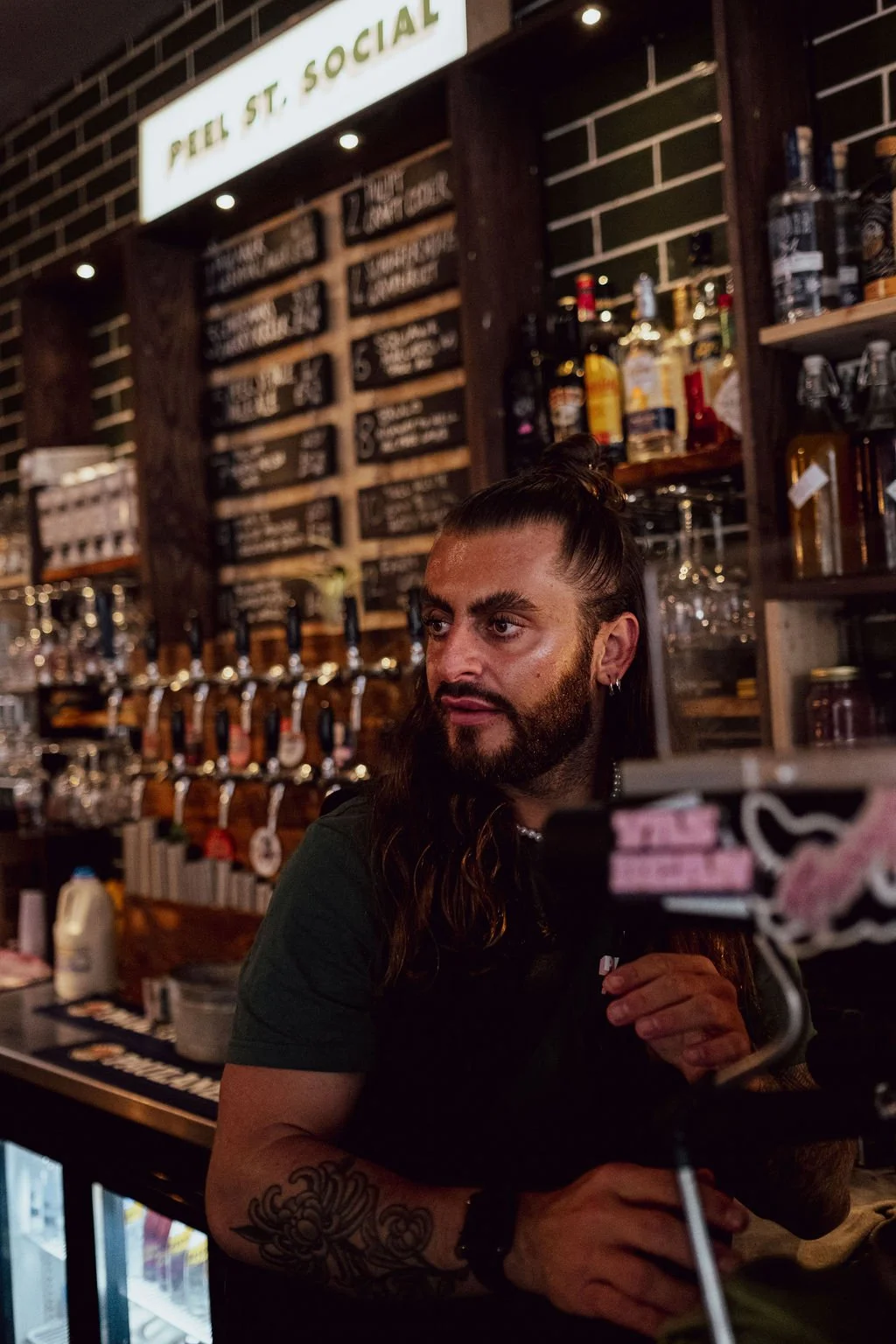 A bartender with long dark hair tied up, a beard, and tattoos on his arm stands behind the bar at a social club or pub, with bottles of alcohol and a chalkboard menu in the background.
