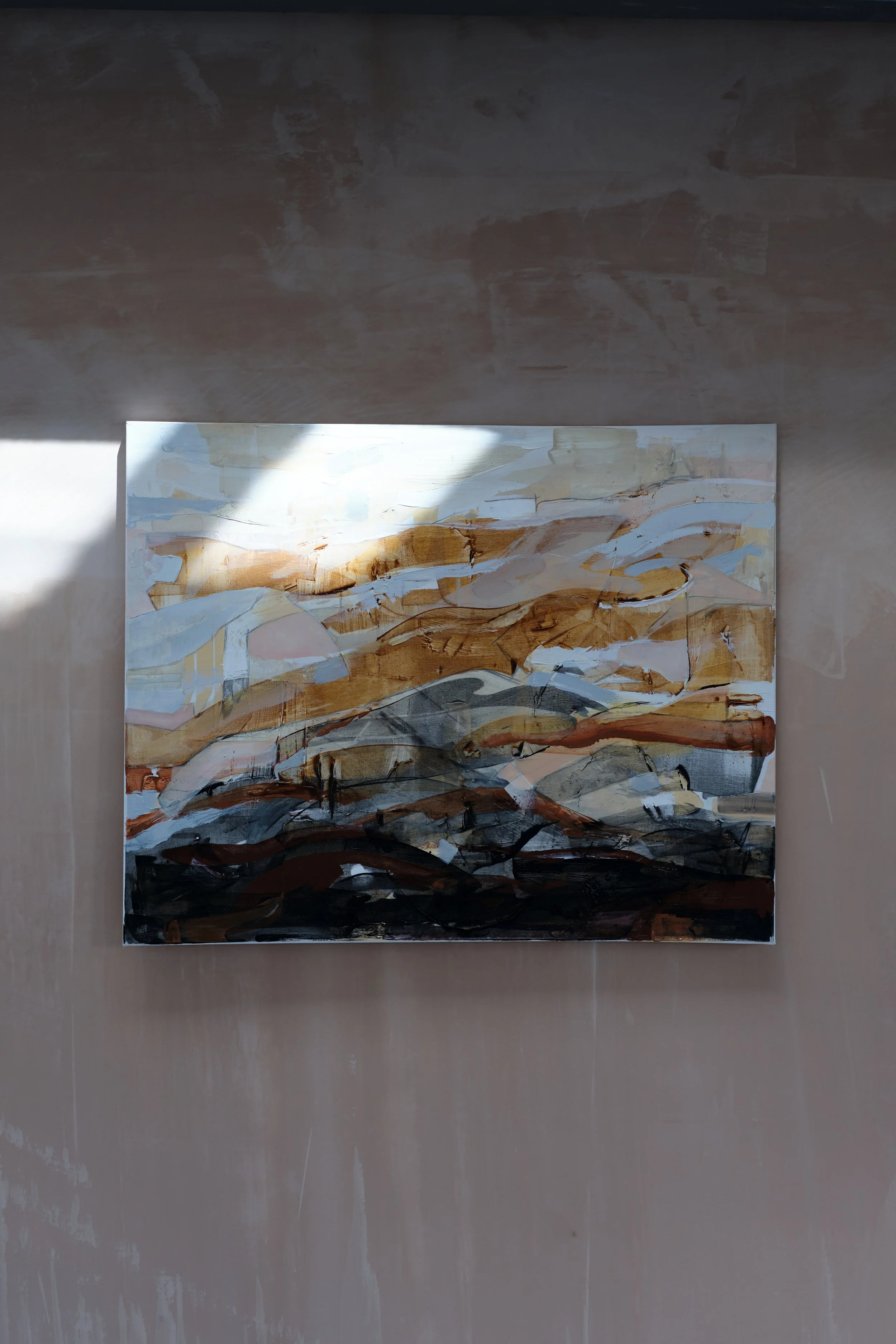 An abstract landscape painting featuring mountain-like shapes in shades of blue, brown, and black, hung on a wall with sunlight casting a shadow on right side.