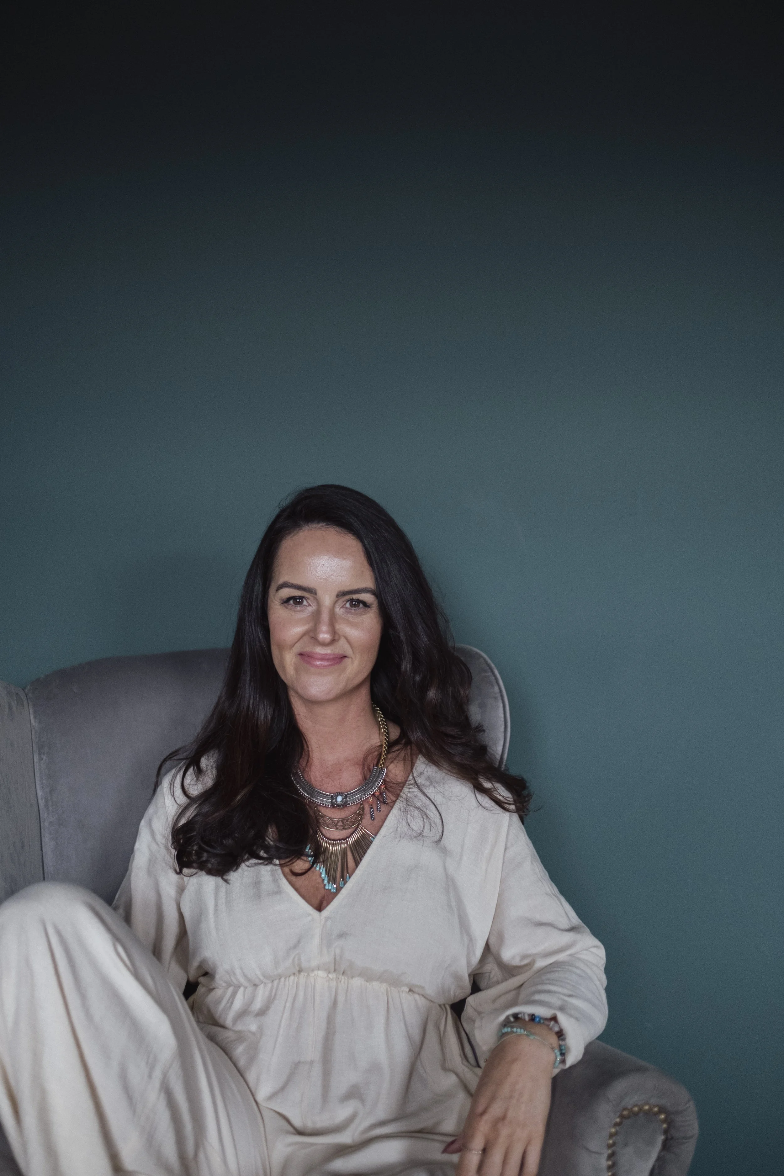 A woman with long dark hair sitting on a gray armchair against a teal wall, wearing a beige dress and layered necklaces, smiling at the camera.