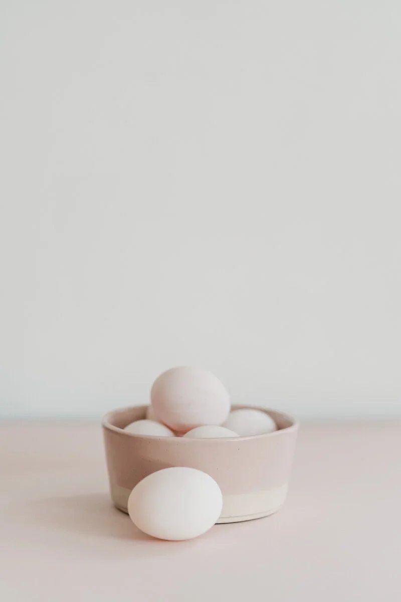 White eggs in a beige bowl with one egg outside the bowl on a light surface against a light background.