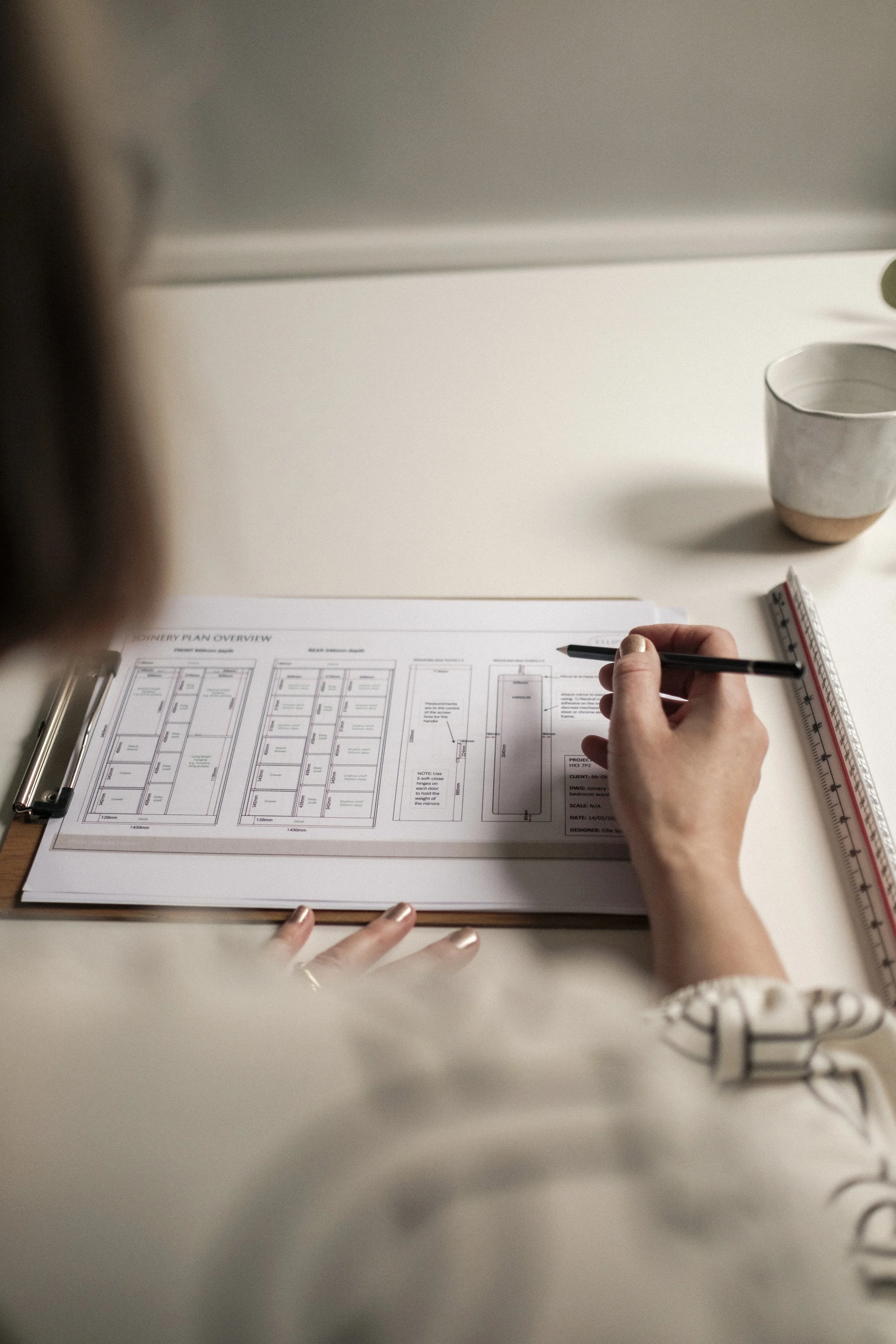 A person is reviewing architectural drawings or plans on a desk, with a pen in hand. There is also a ruler, a small potted plant, and a cup of water.