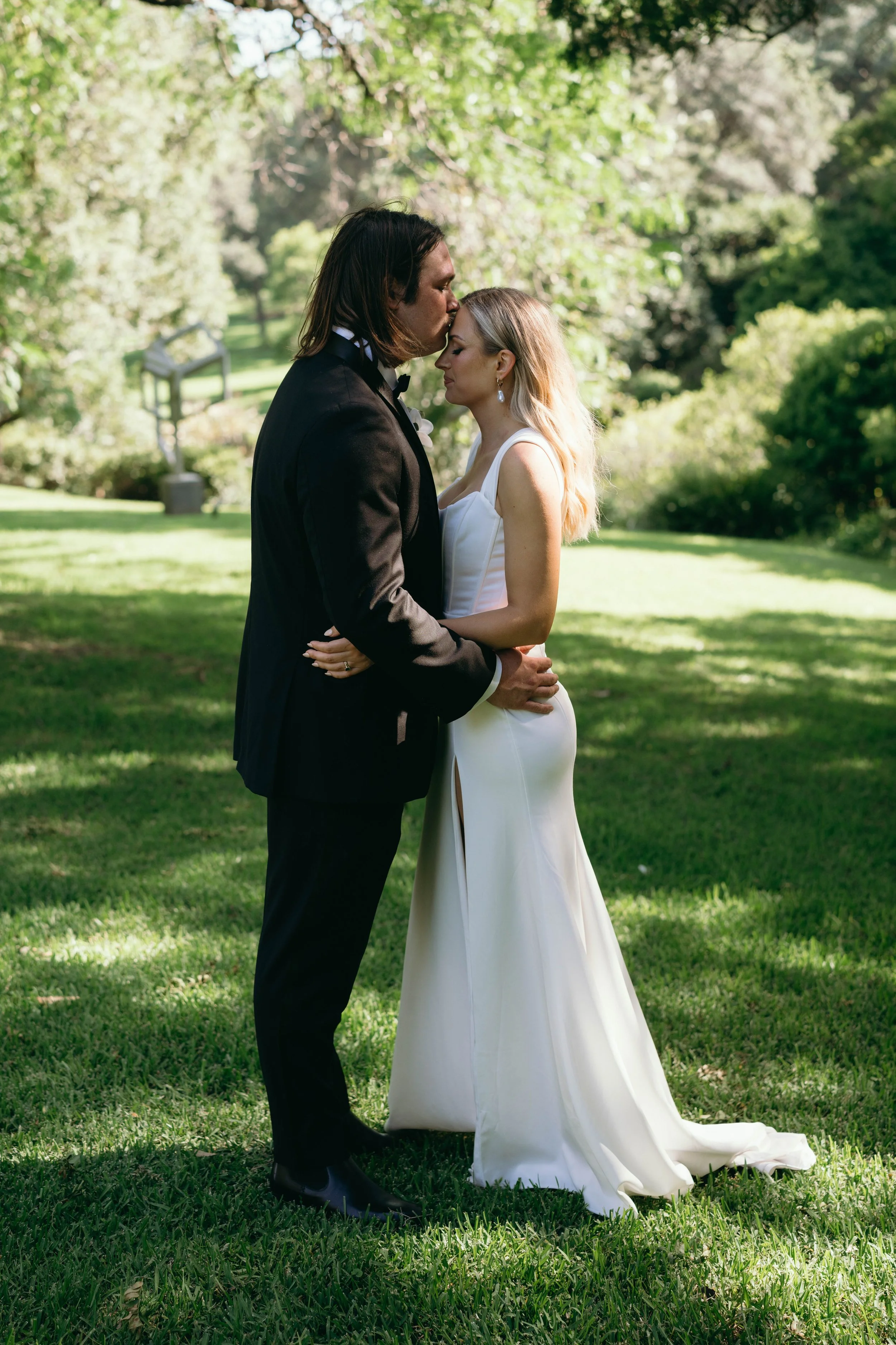 A couple in wedding attire sharing a tender moment outdoors, surrounded by green trees and grass.