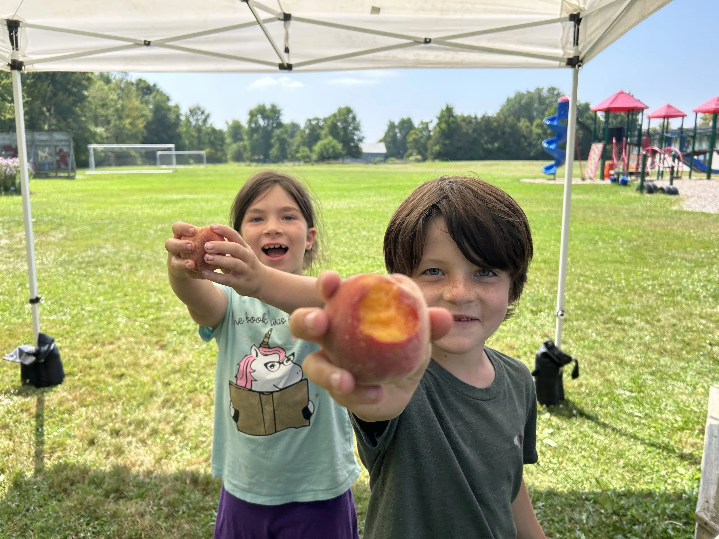 2 smiling campers, holding peaches up to the camera