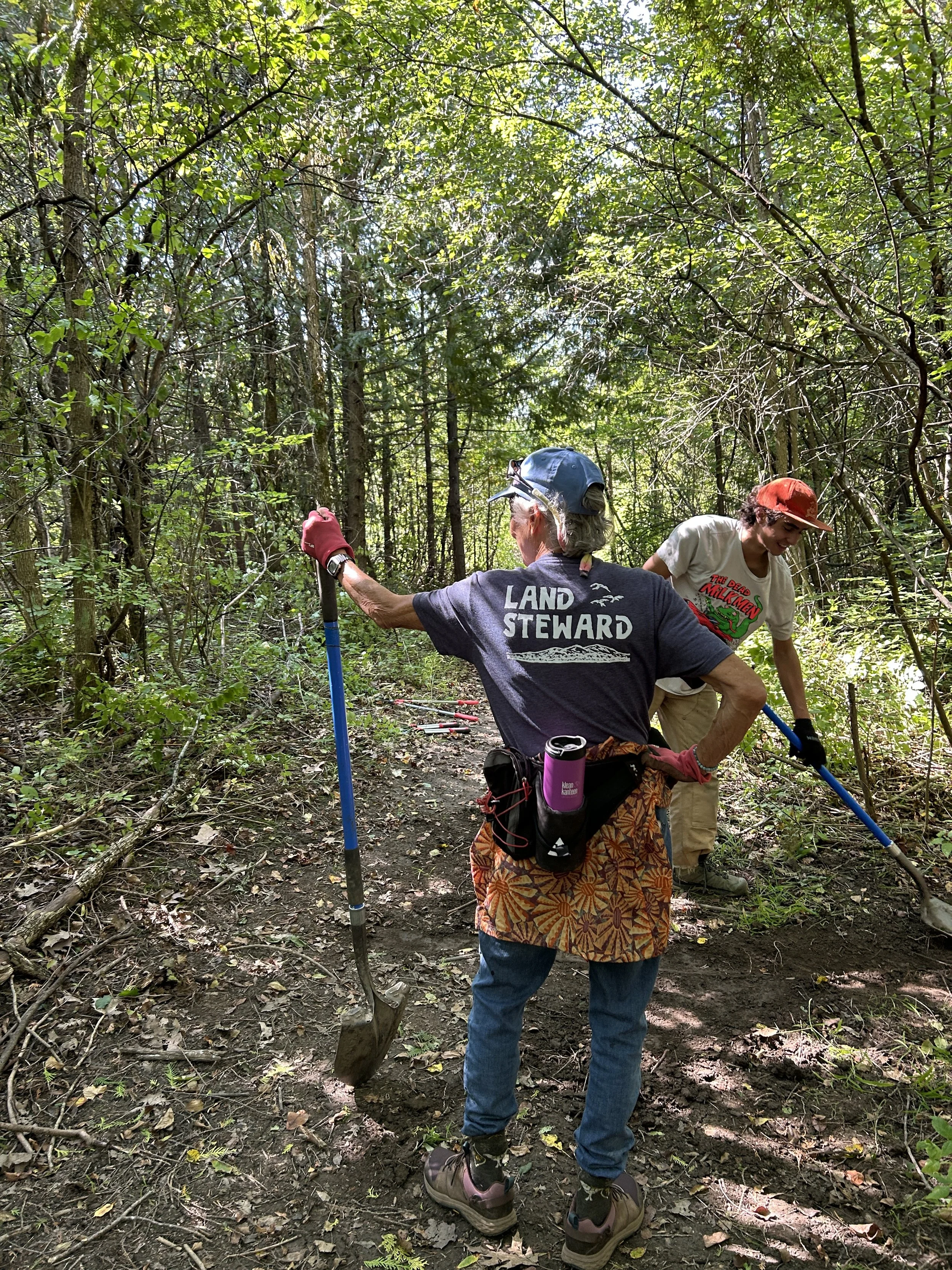 two volunteers working on a dirt trail. One stands with her back to the camera, modeling the Land Stewards shirt
