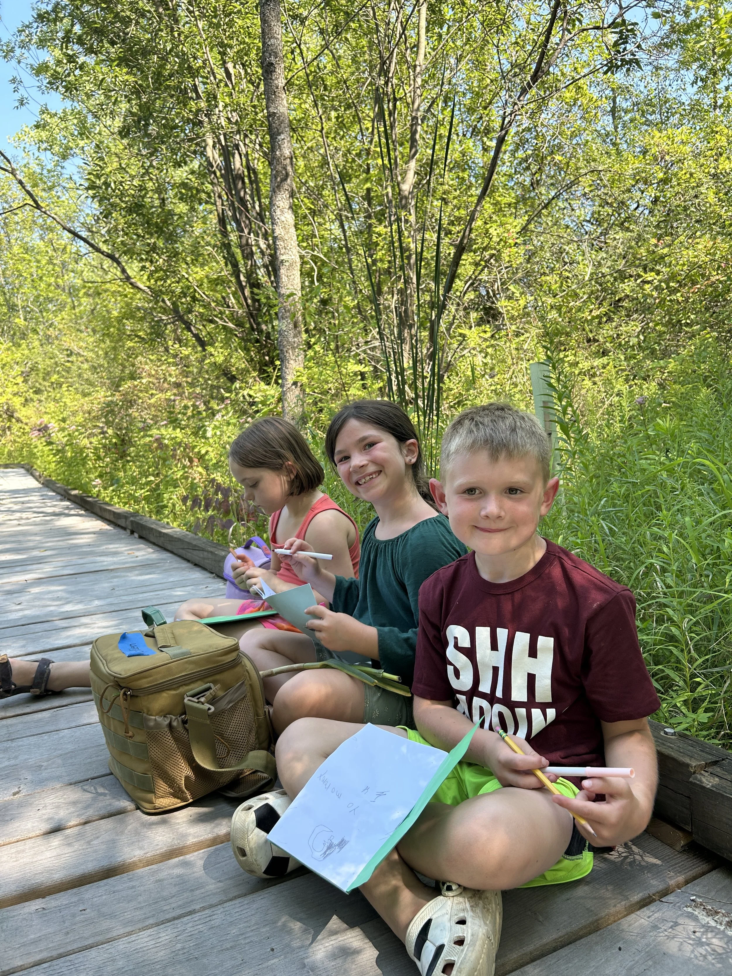 3 campers on the Rec Park boardwalk, working on their nature journals