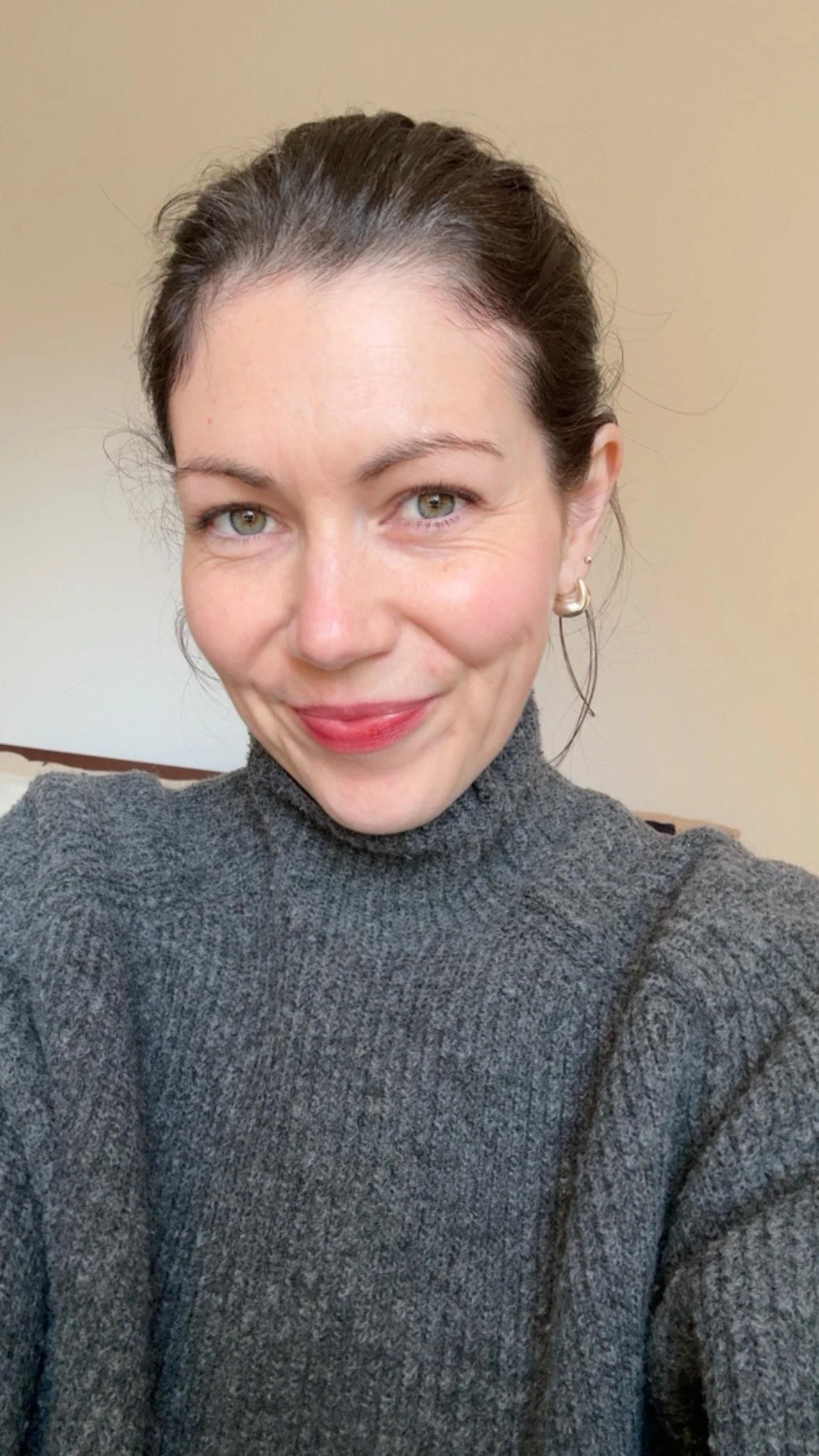 A woman with dark hair tied back, wearing a gray turtleneck sweater and hoop earrings, smiling in a well-lit room with a beige wall in the background.