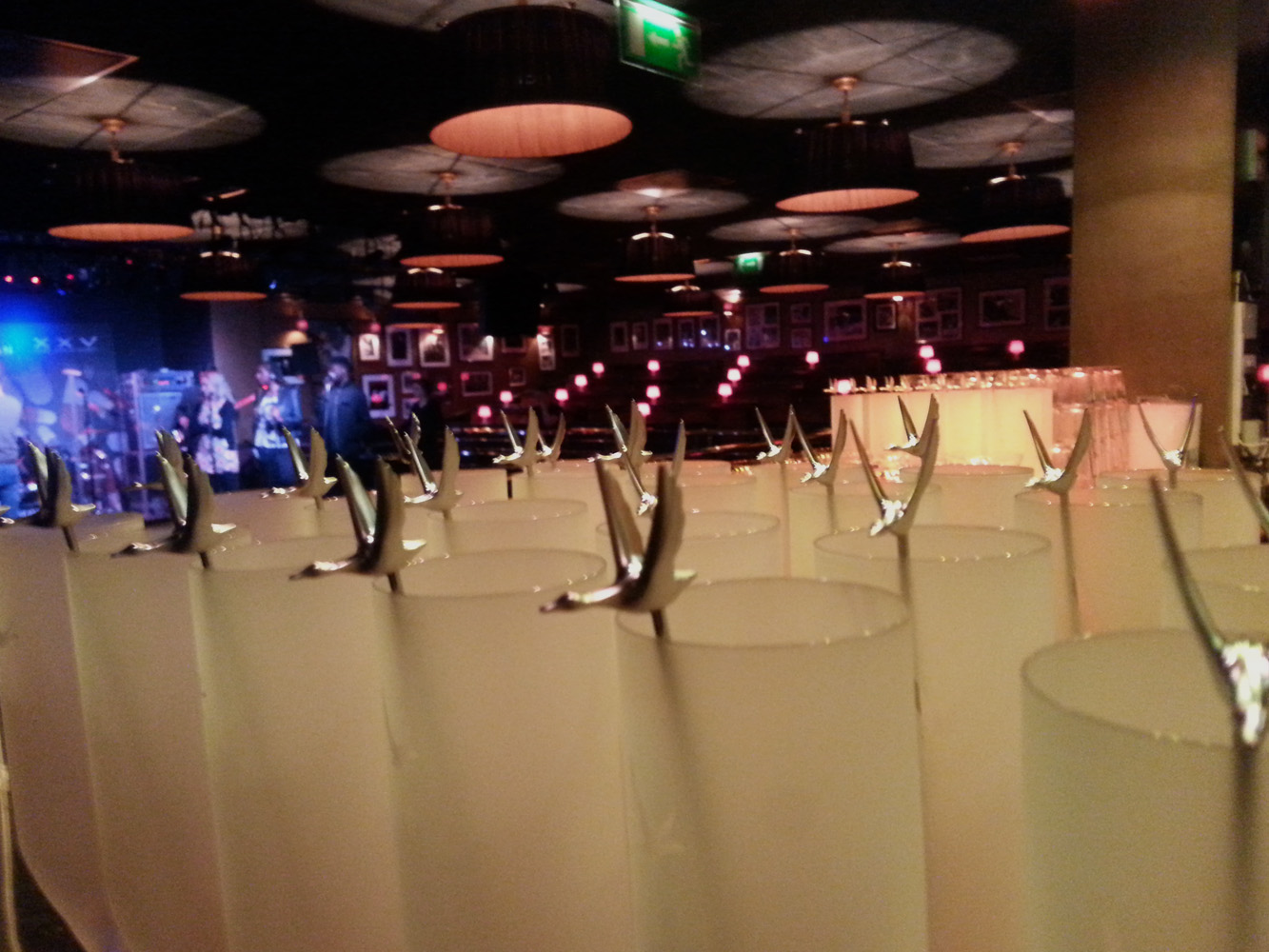 Empty tables with white cups and silver spoons arranged upside down in a row in the foreground, a stage with musical instruments and performers in the background, dim lighting with pink and blue accents, and a dark ceiling with hanging light fixtures