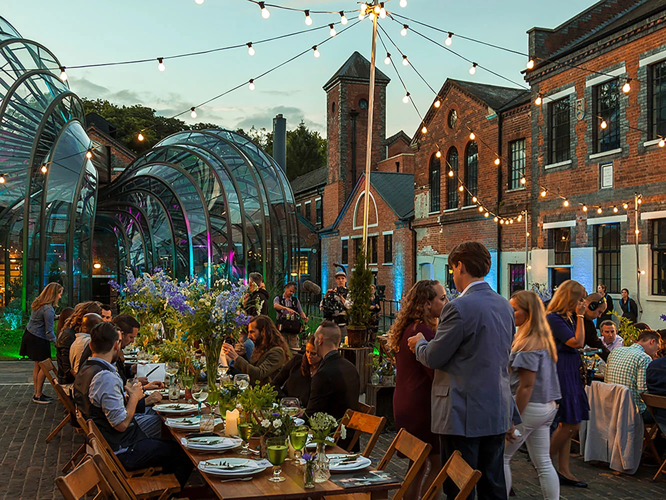 Outdoor evening event with people socializing and dining, decorated with string lights, large floral centerpieces, and a glass greenhouse structure, set against brick buildings.