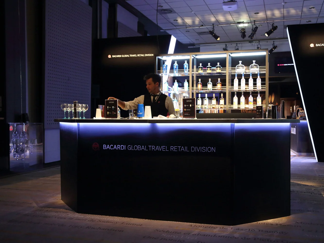 A bartender stands behind a bar counter with bottles of alcohol displayed on illuminated shelves behind him. The bar has a sign reading "Bacardi Global Travel Retail Division." The bartender prepares drinks in a dimly lit environment with stage light