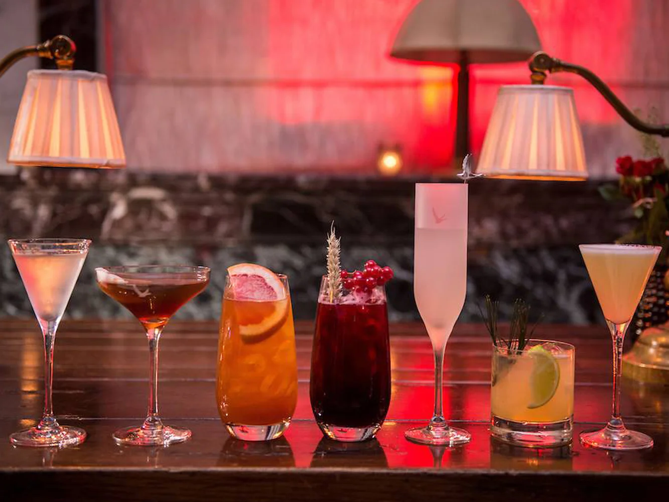 Six colorful cocktails on a wooden bar with warm lighting and lamps in the background.