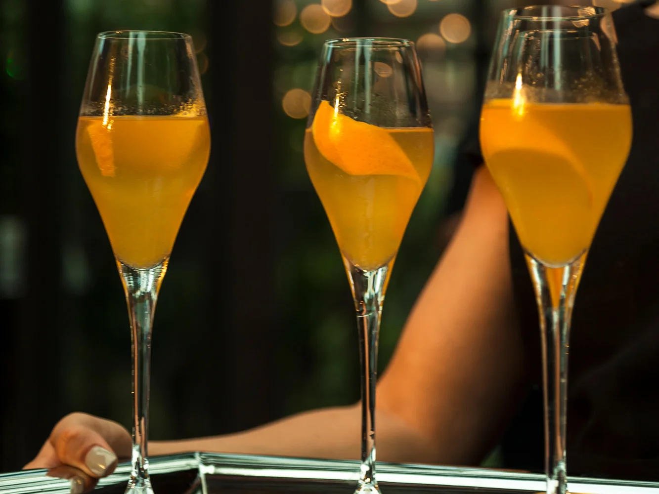 Three champagne flutes filled with yellow-orange mimosa drinks, garnished with orange slices, held on a silver tray.