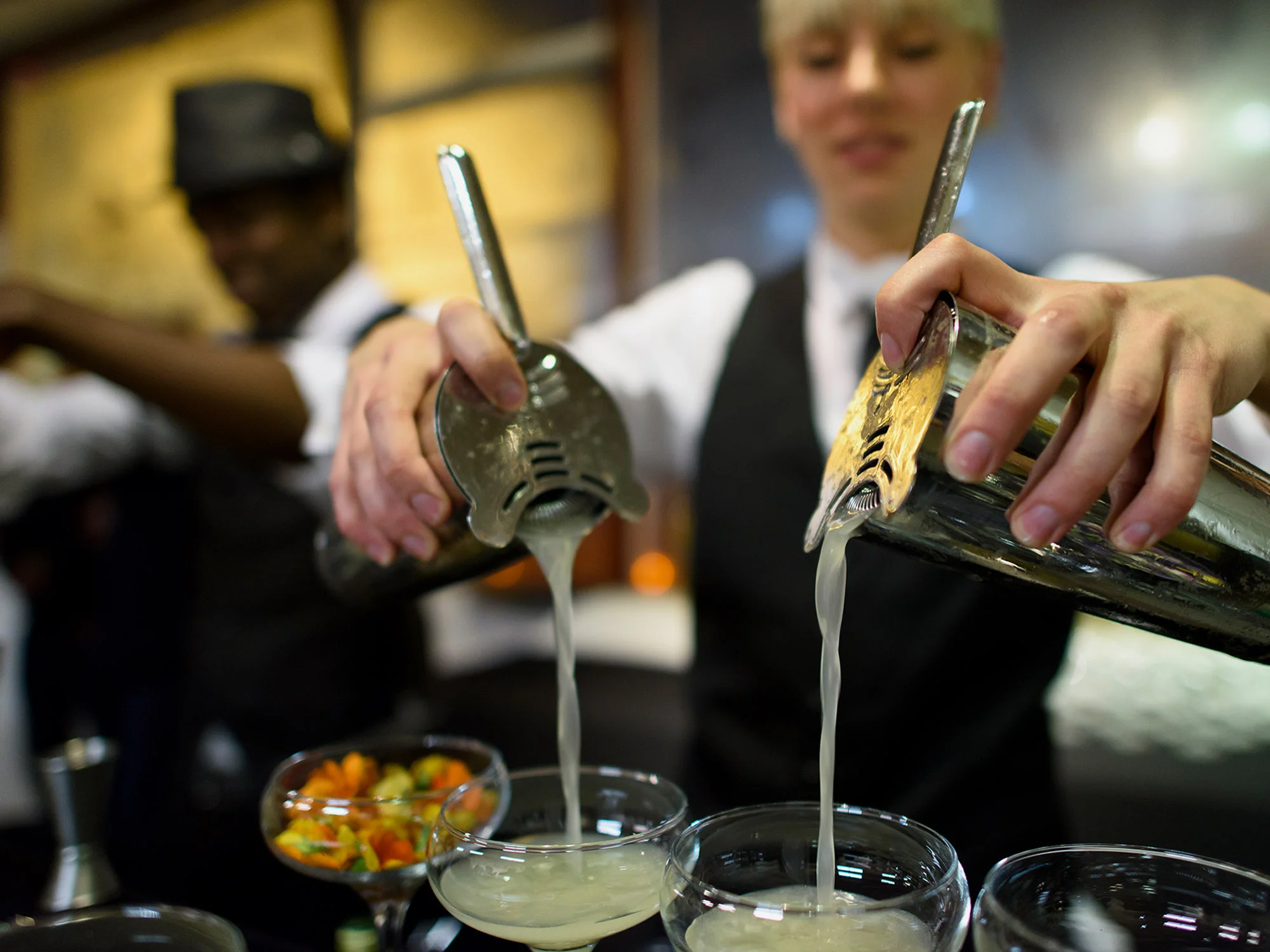 Bartenders pouring drinks into glasses at a bar, with a bowl of colorful snacks called 'pico de gallo' in the background.