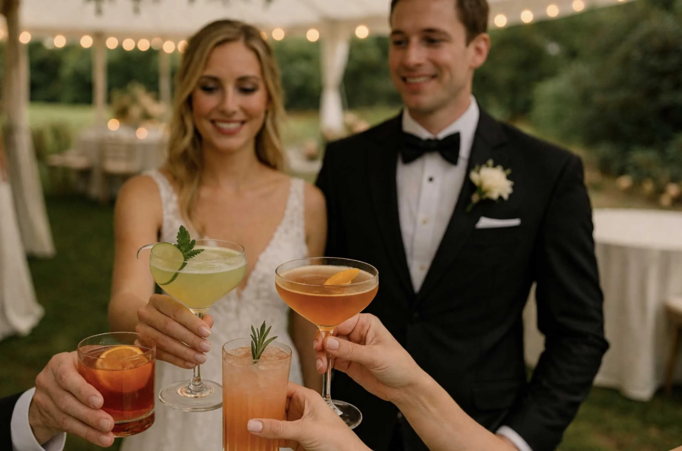 Bride and groom in formal wedding attire toasting with colorful cocktails at outdoor wedding reception.