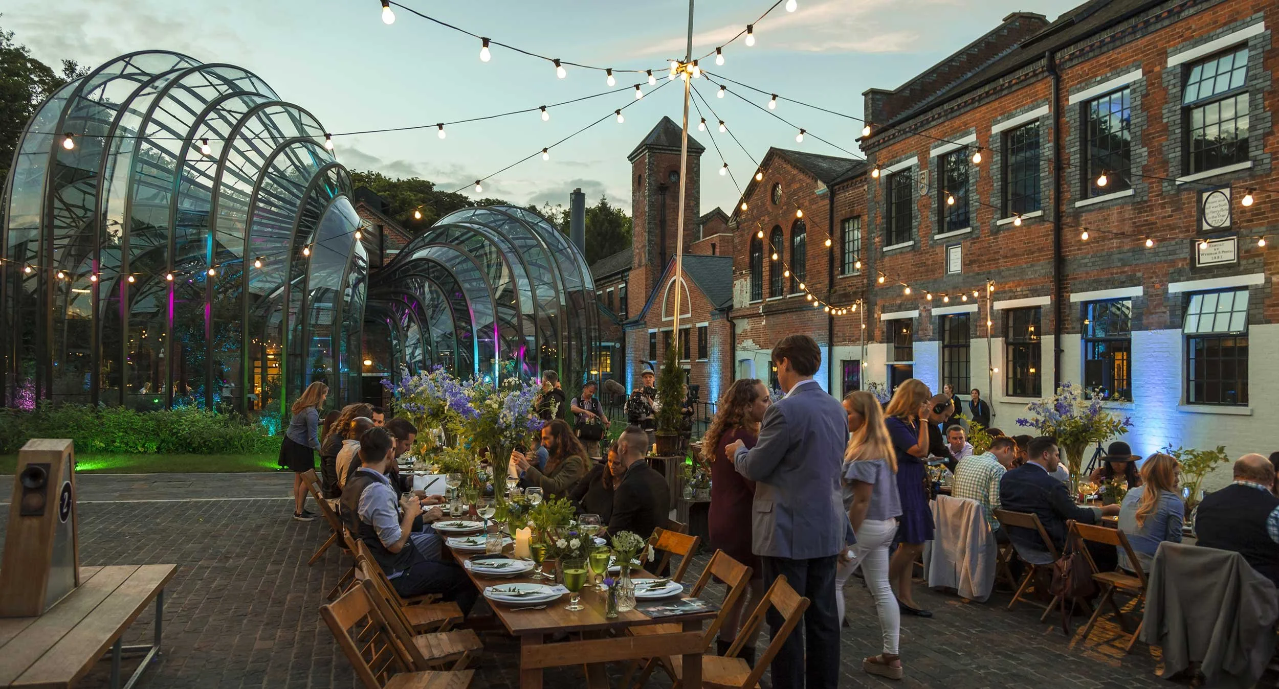 An outdoor party at dusk with people dining and socializing, decorated with string lights. There are curved glass greenhouses and brick buildings in the background, with floral arrangements on the tables.