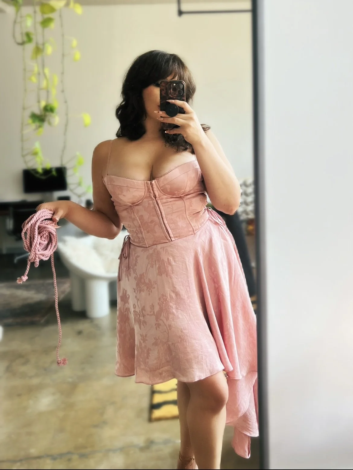 Woman taking a mirror selfie wearing a pink strapless dress with a corset bodice, holding pink rope, in a room with a white chair, a black table, and a plant in the background.