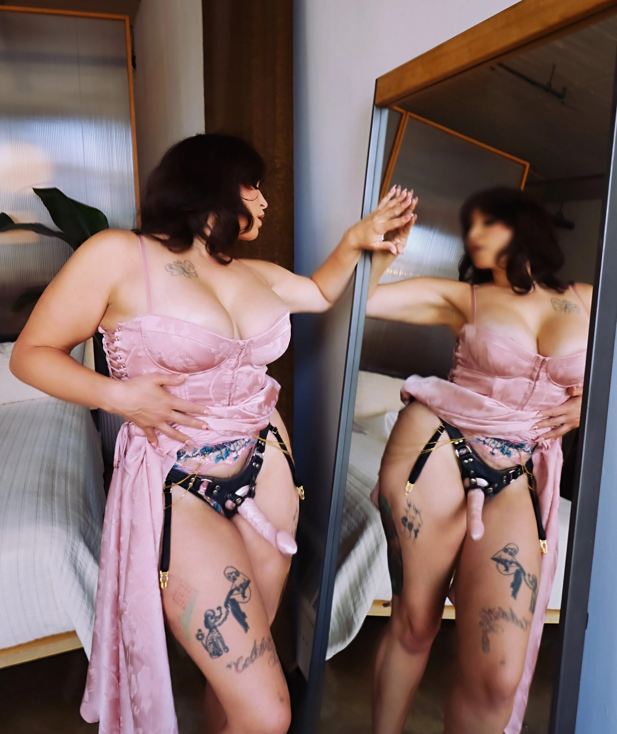 Gentle femdom Zoey Belladonna stands in front of a mirror in a pink dress wearing a pink strap-on that is peeking out from under her skirt.