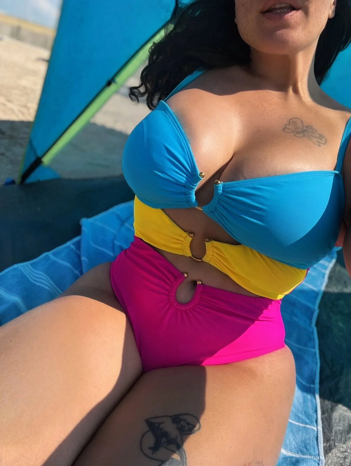 Close-up of a woman wearing a colorful swimsuit at the beach, with tattoos visible on her chest and thigh.