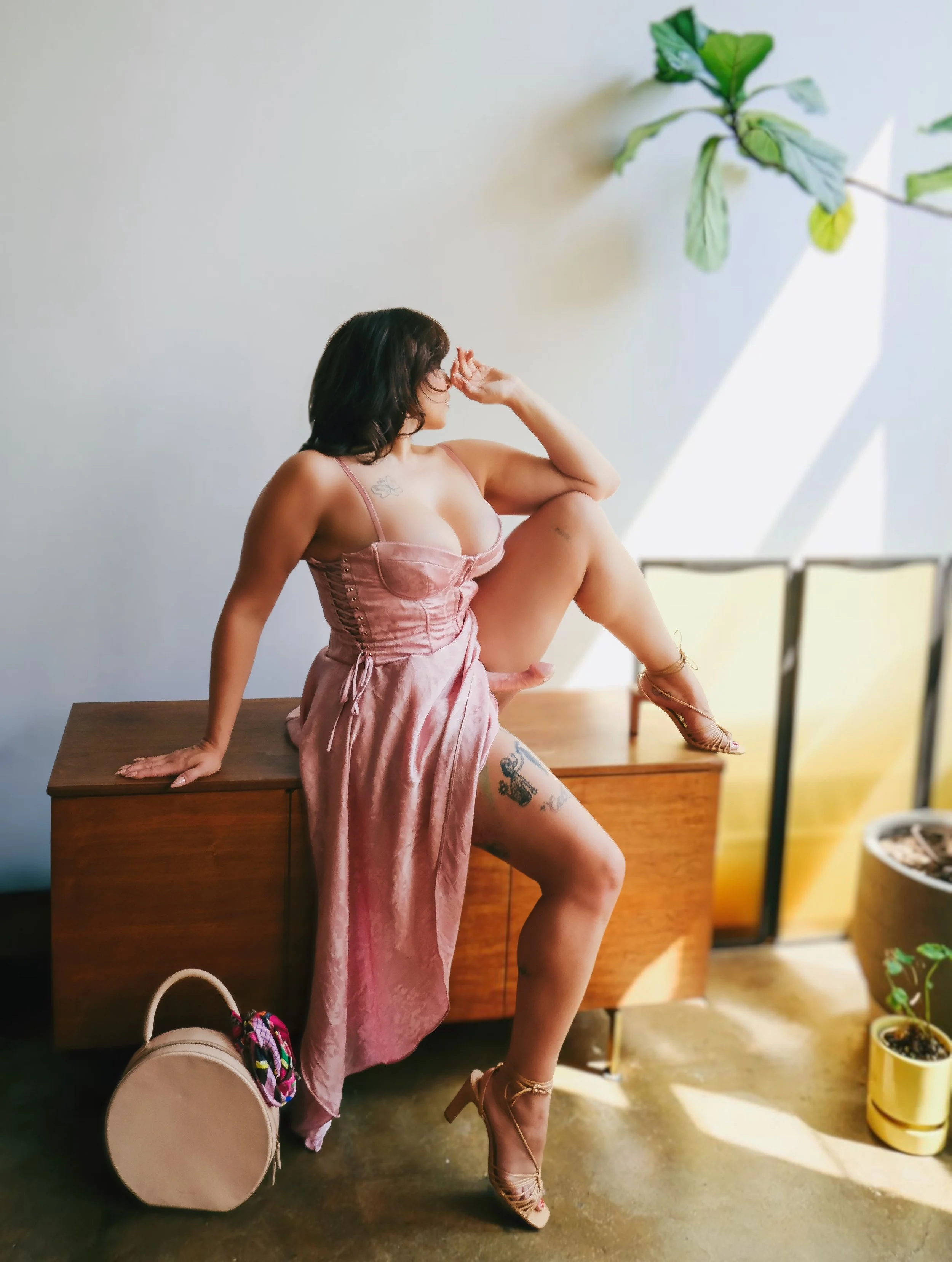 Gentle femdom Zoey Belladonna sits atop a dresser in a pink dress wearing a pink strap-on that is peeking out from under her skirt.