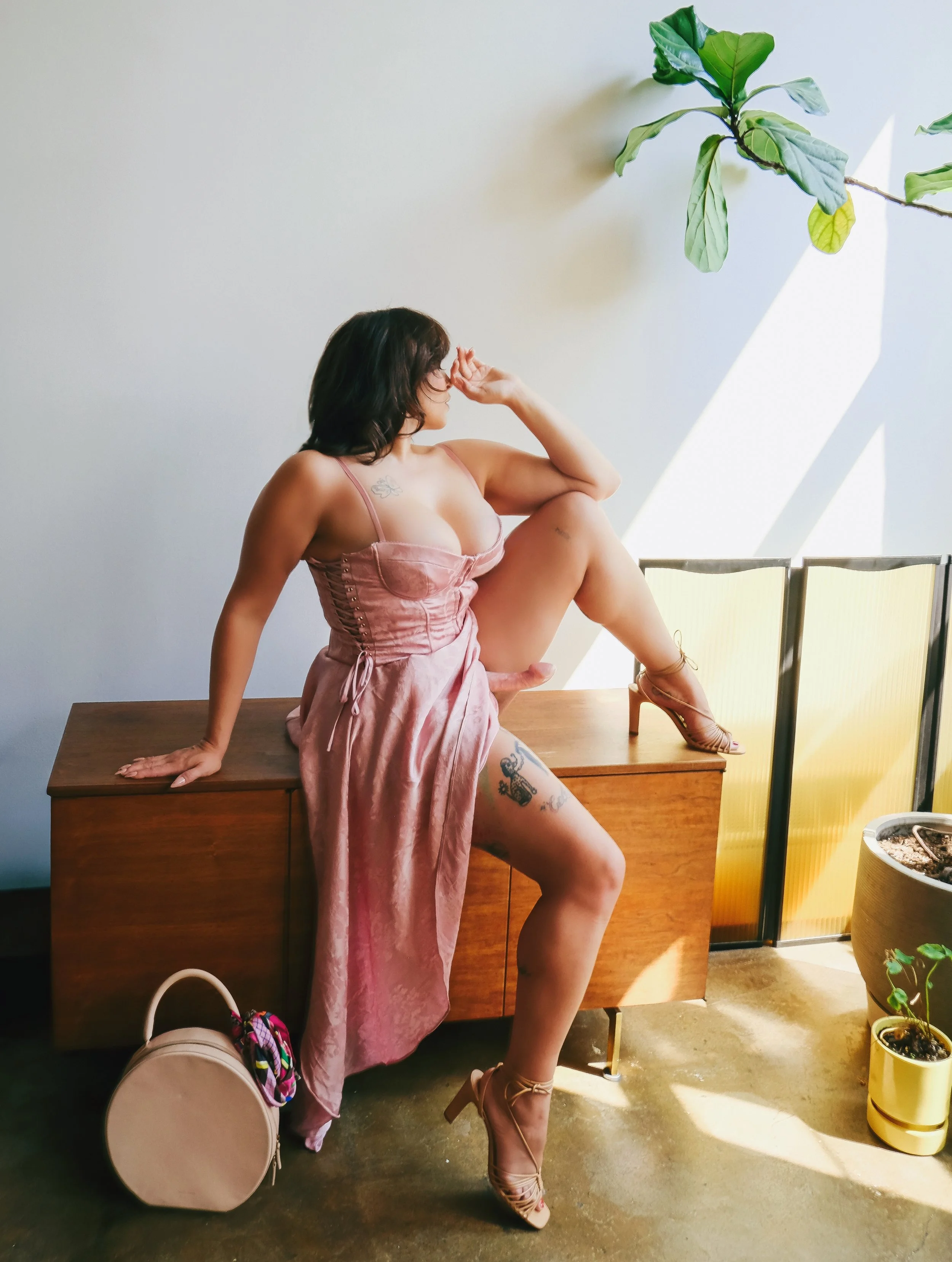 Gentle femdom Zoey Belladonna sits atop a dresser in a pink dress wearing a pink strap-on that is peeking out from under her skirt.