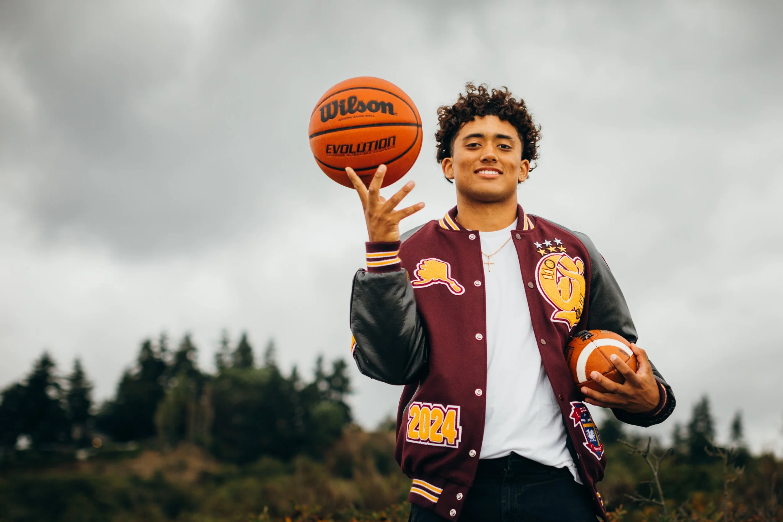 black senior in letterman jacket holding basketball and football
