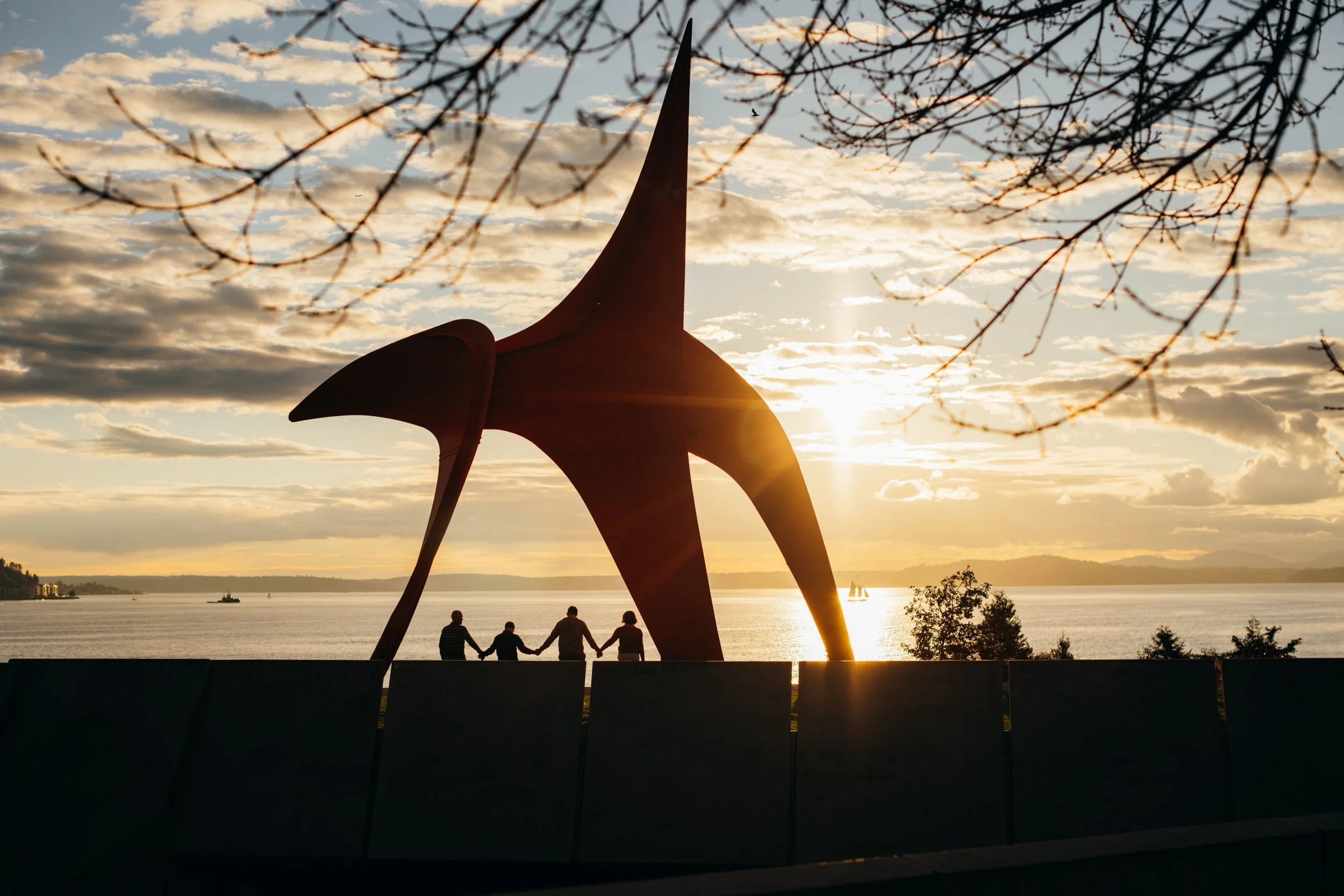 An artistic image of a family of 4 holding hands underneath a giant sculpture at the Olympic sculpture park in Seattle.  The sun is setting and they are pictured from far away in silhouette.   