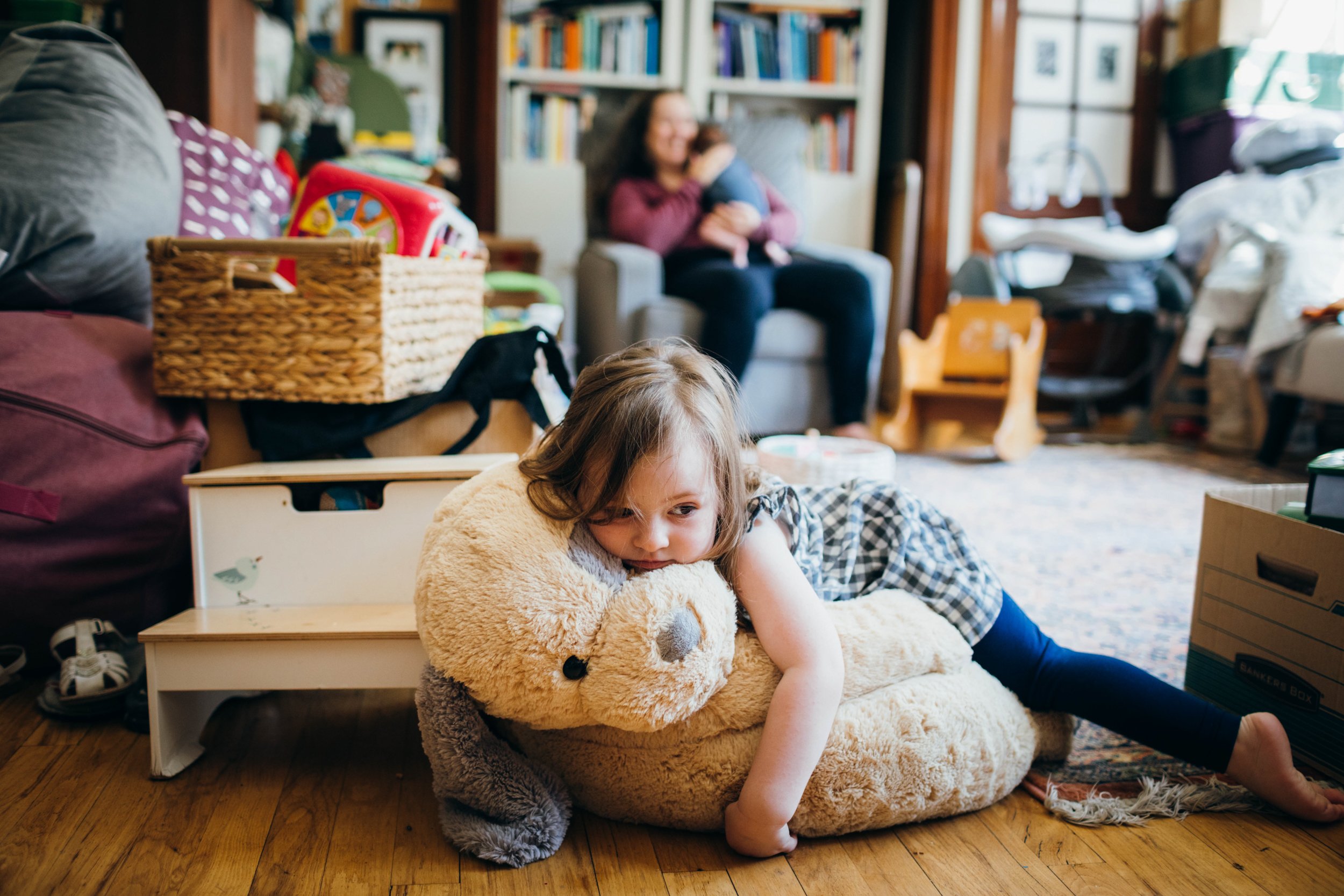  A bored toddler lays on her giant puppy chair as her Mom holds her baby brother in the background.  They are at their home in West Seattle.   