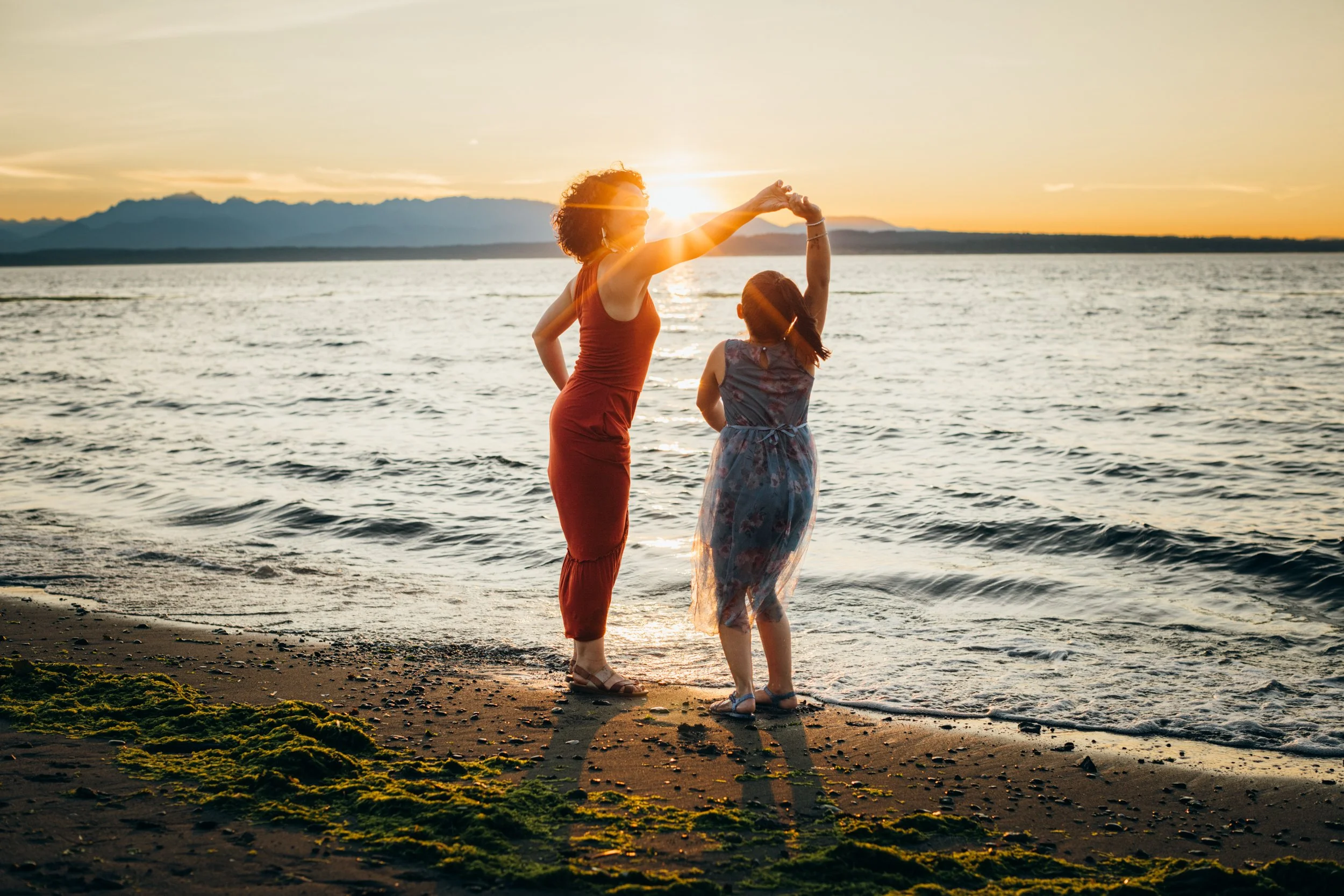  A joyful shot of a mother and daughter dancing in the water with the sun setting behind the mountains at sunset.  