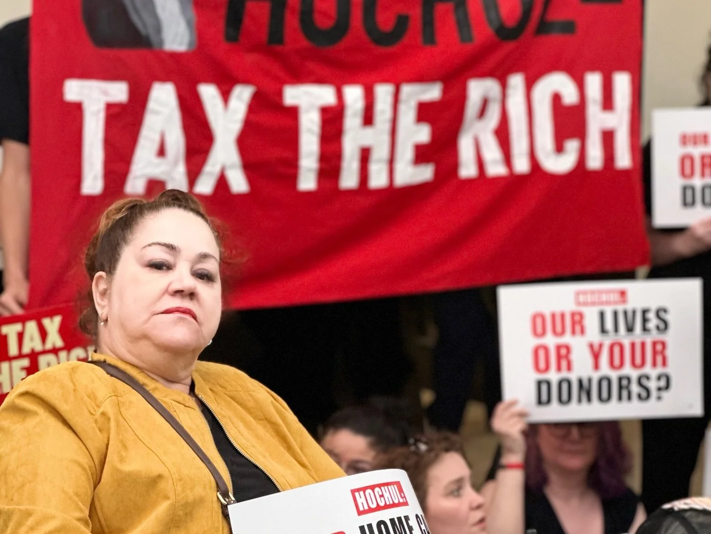 An unsmiling woman stares into the camera with a tax the rich banner behind and people holding signs reading Hochul: Our lives or your donors?