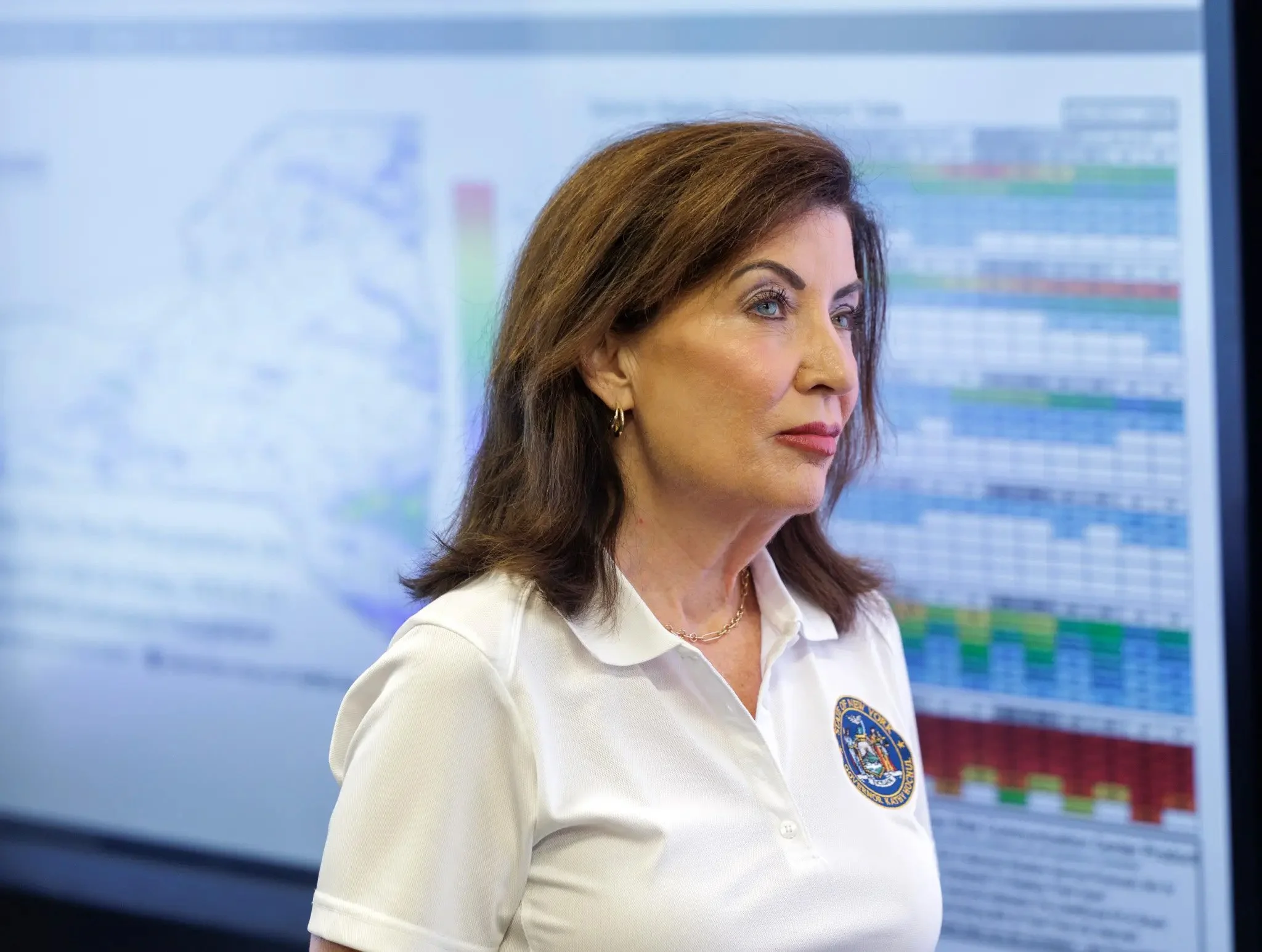 Kathy Hochul in a white shirt looks off to the right of the image. Behind her are large charts which are mostly blurry and blue from way the image was shot