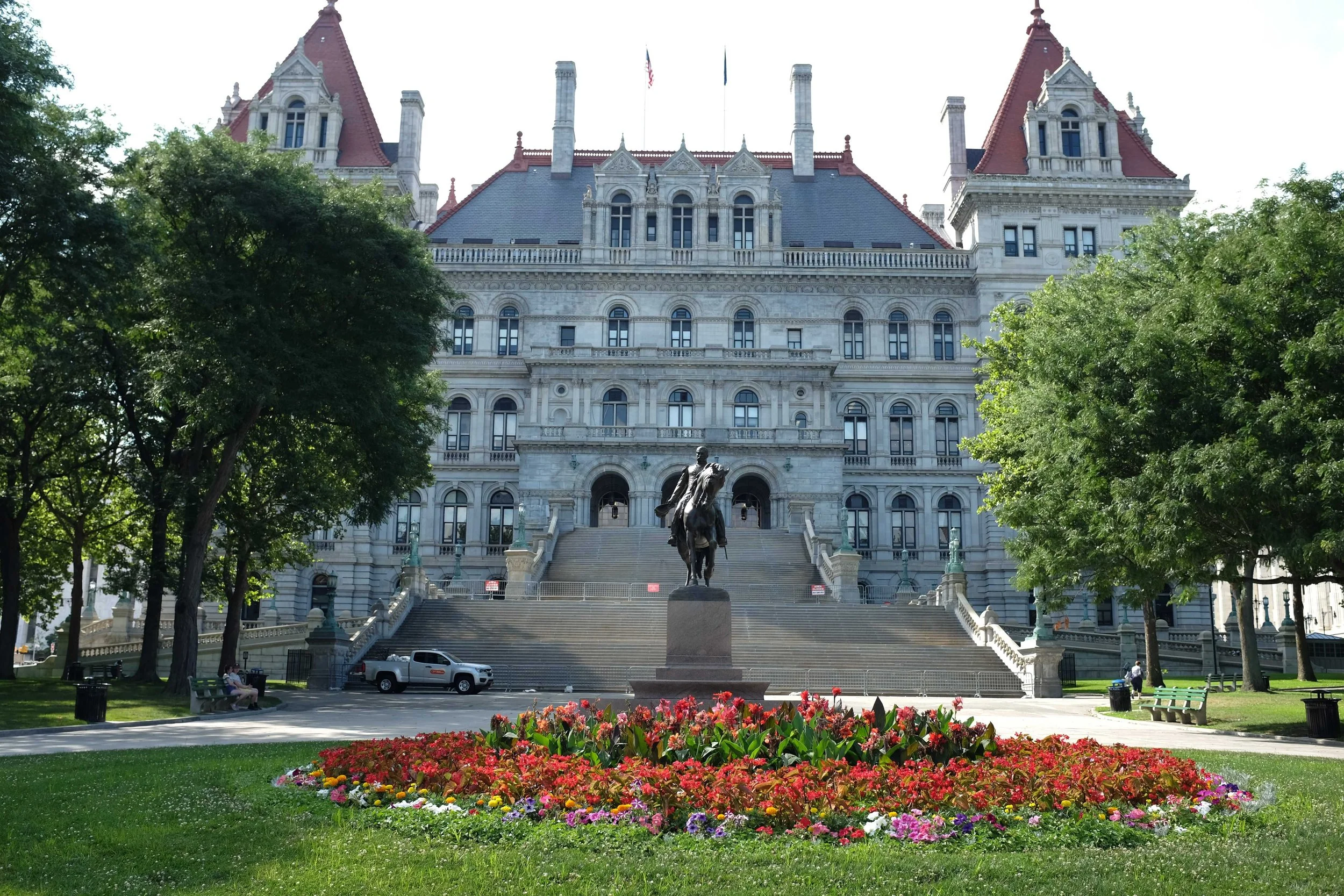 View of the capital building with stairs coming down in front of it, a garden of flowers in the foreground, a sculpture of a man on a horse above the flowers, and trees framing the street. A grey pickup truck sits at the base of the stairs.