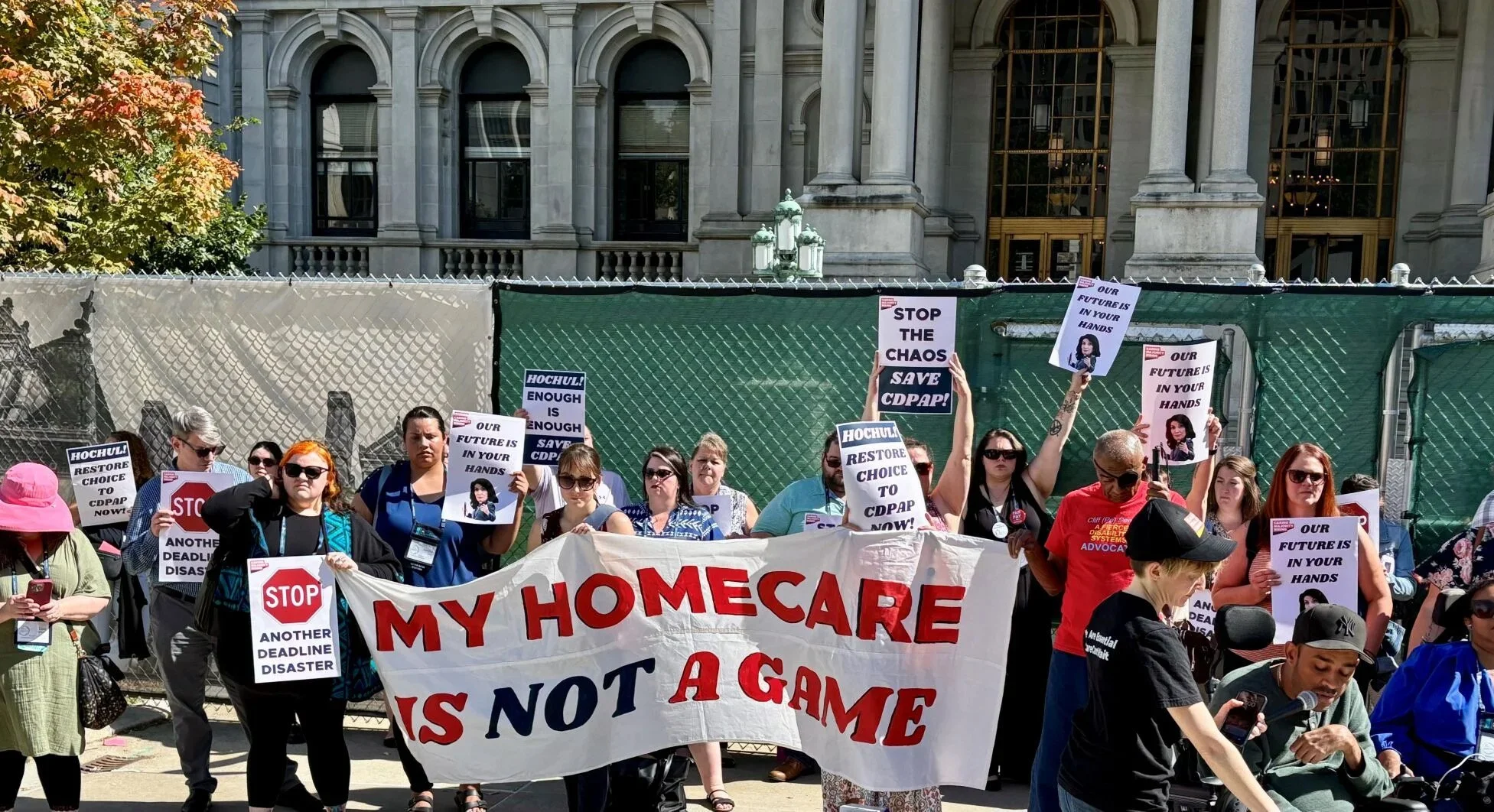 About 20 protestors stand outside in front of the capital building holding signs and a banner that reads "My Homecare Is Not a Game"