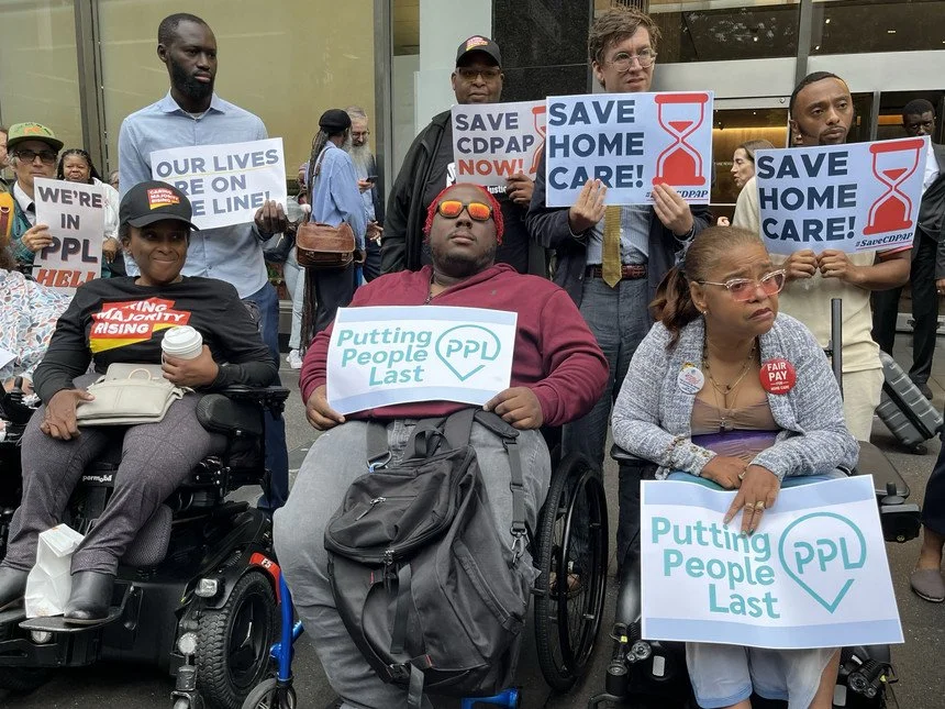 Eight protests sit and stand with signs reading PPL - Putting People Last and Same Home Care. One person wears a Caring Majority Rising shirt and baseball cap. Others are visible in the background.