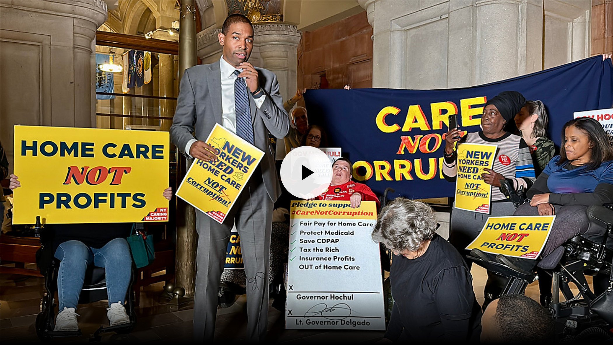 Lt. Gov. Antonio Delgado speaks into a microphone holding a sign reading New Yorkers Need Home Care. There are a small group of disability advocates standing behind him holding signs and a banner.