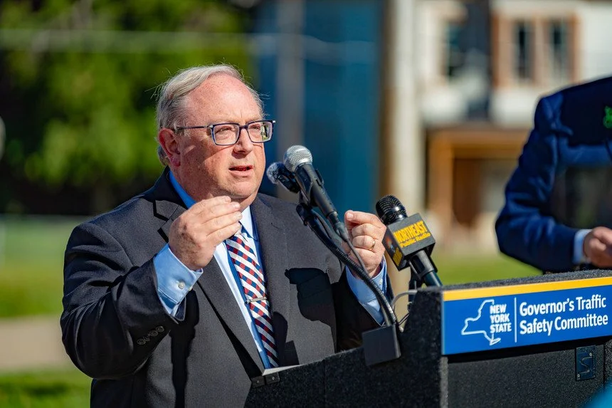 Health Commissioner James McDonald stands at a lecturn with microphones, speaking and gesturing with his hand. He is in a suit. The lecturn reads New York State Governor's Traffic Safety Committee