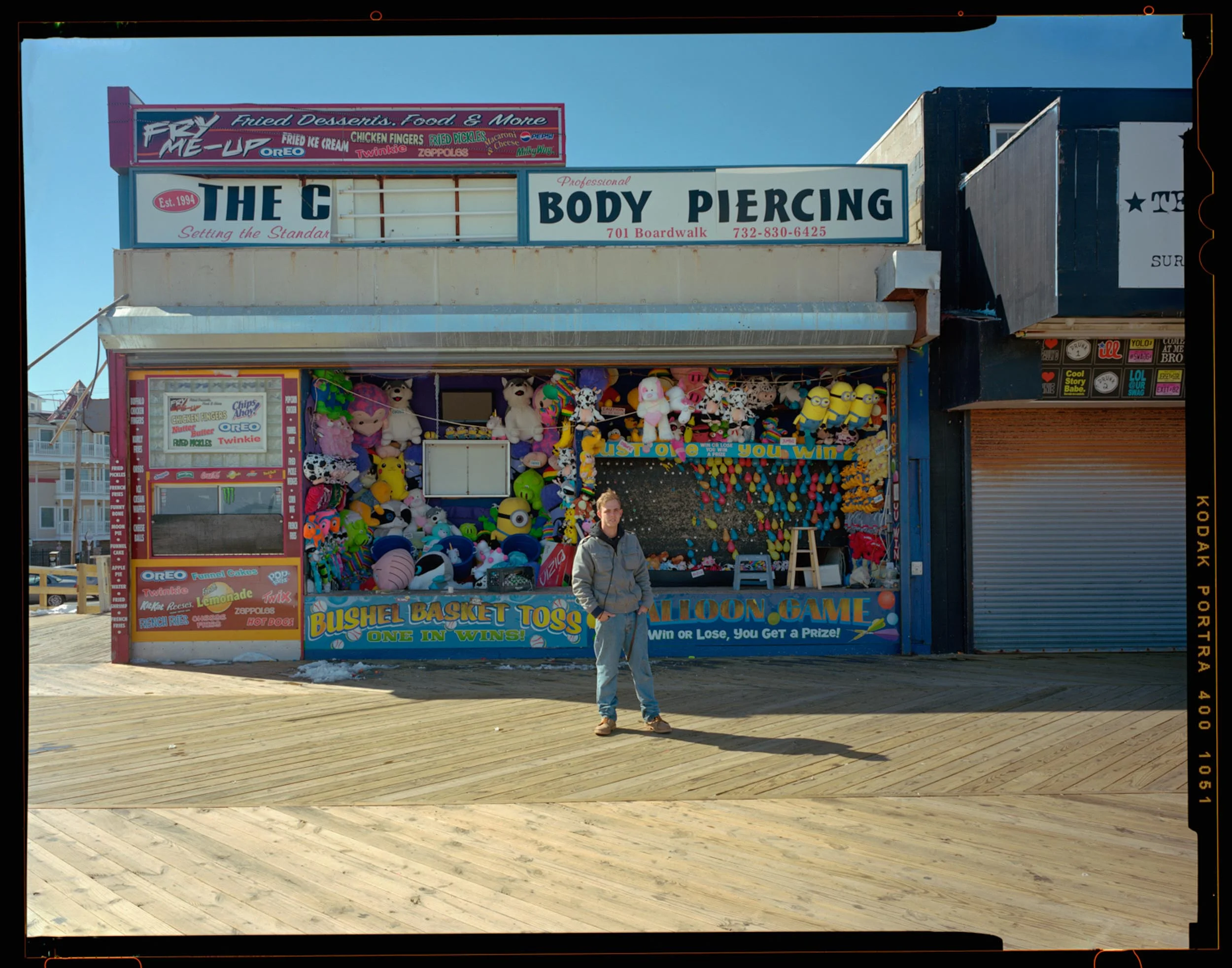Body Piercing, Seaside, NJ. 2014. 4x5 Shen Hao It was a bright sunny cold day as I was walking down the boardwalk with my 4x5 camera on my shoulder. I said hello and the man at the shop took interest in my camera. After talking and he told me he had interest in photography so I showed him how the camera worked. He eventually asked me what I was doing, so I explained and asked if he would mind standing for a portrait. He was hesitant at first so I pulled back and tried to capture the whole place along with him so he would feel more comfortable. I actually never caught his name. Nice Kid.