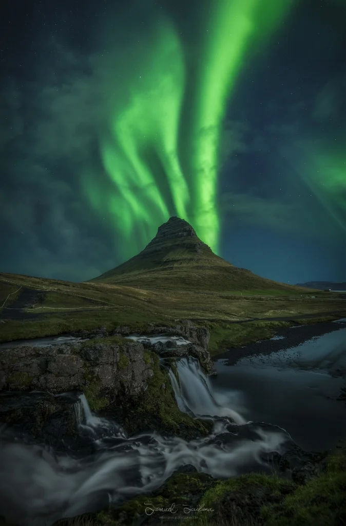 Aurora bands dance above Kirkjufell with Kirkjufellsfoss waterfall in the foreground.
