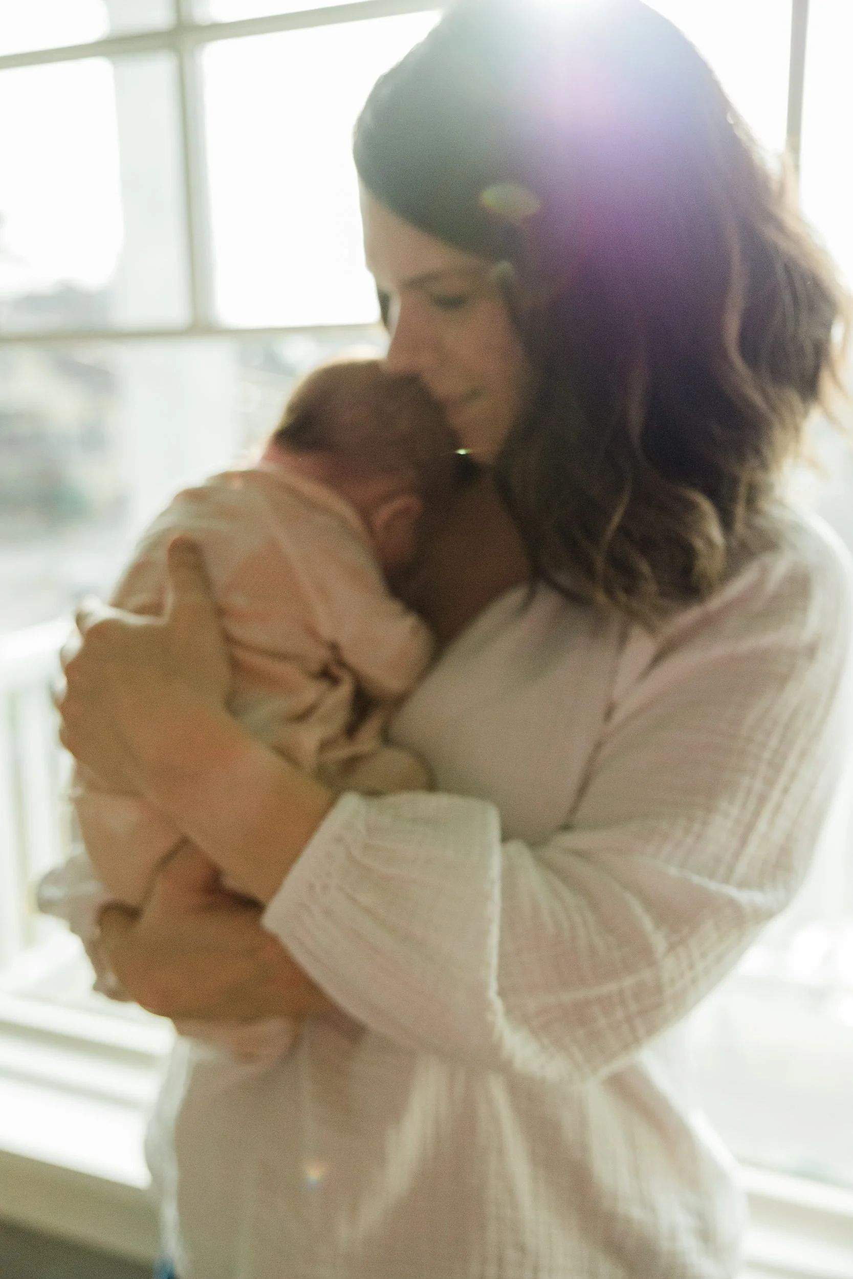 mom cradles baby against her chest while standing in front of window