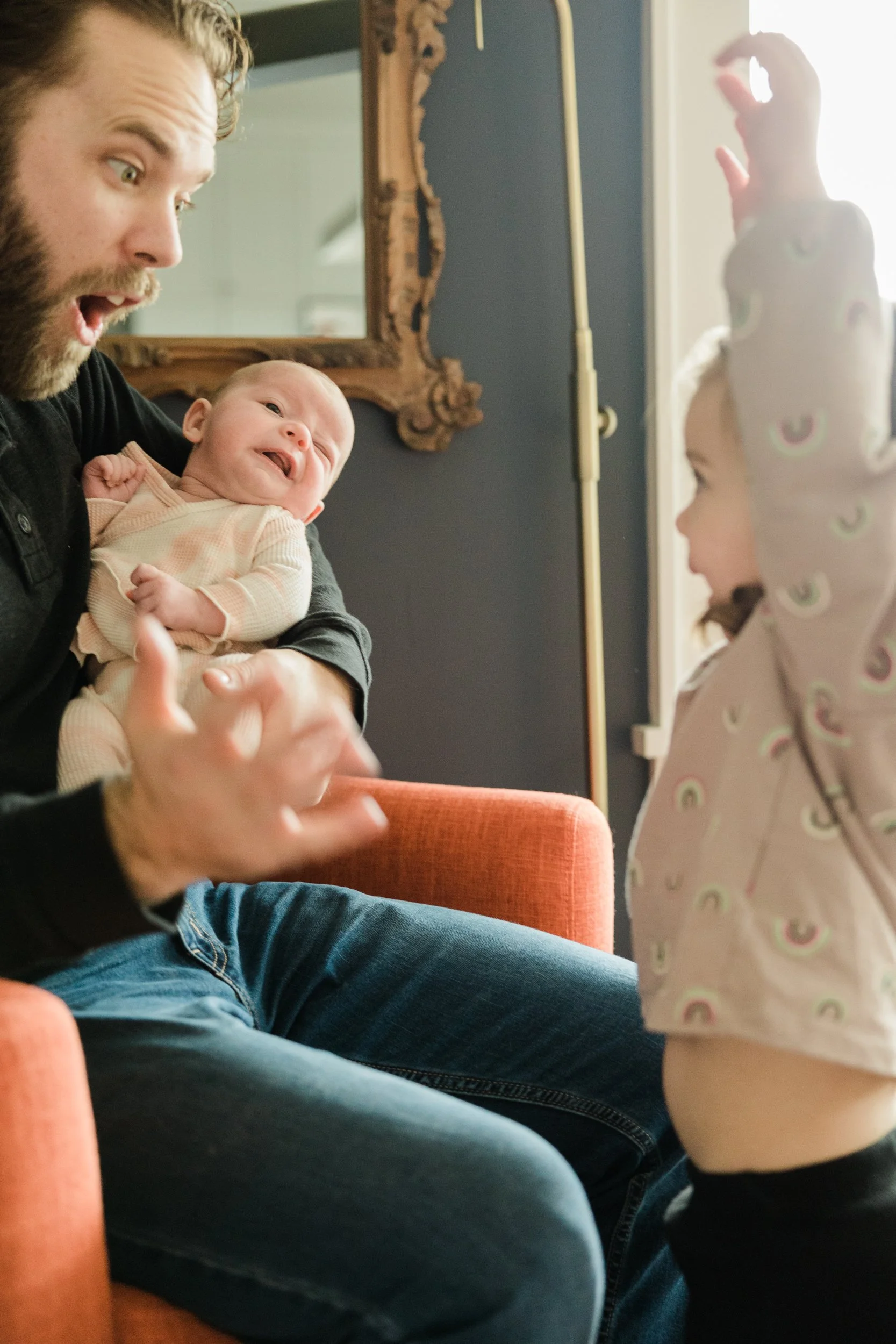 dad sits in chair holding baby while making a surprised face at small girl standing in front of him