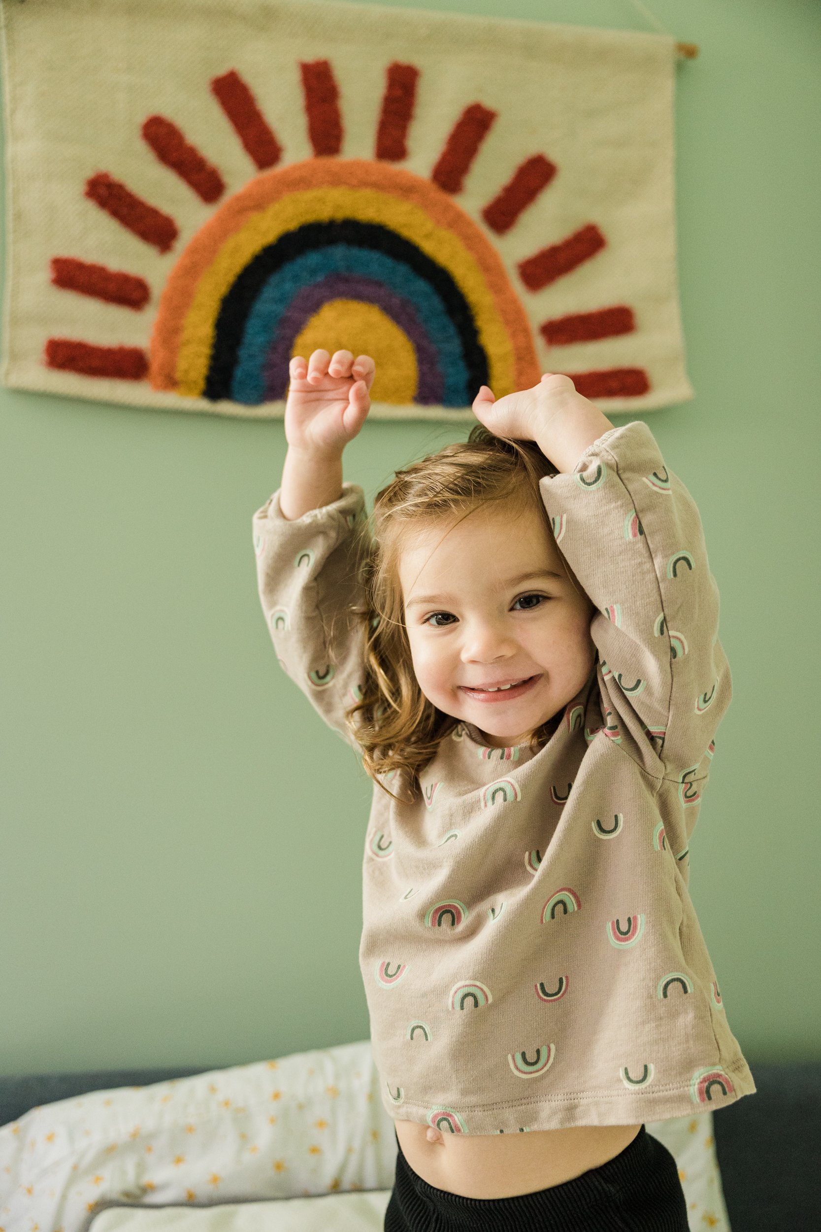 little girl raises her arms above her head with rainbow art on the wall behind her 