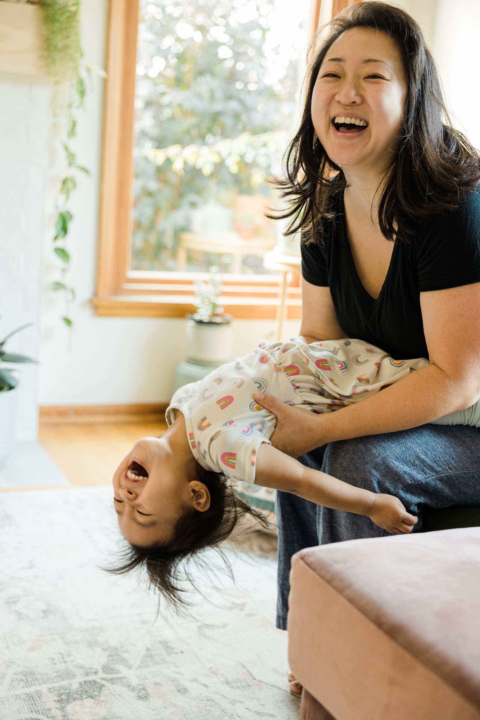 mother sits on couch and daughter flops back over her knees laughing