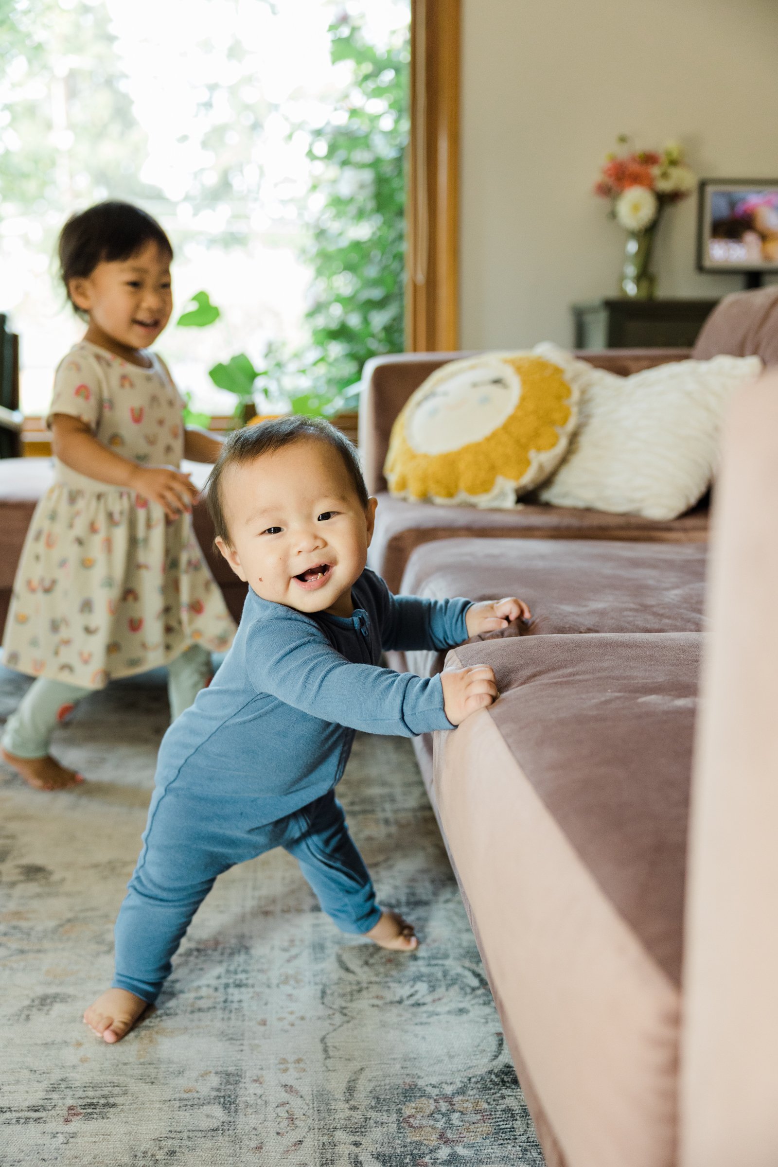 infant stands on ground holding onto pink couch while smiling