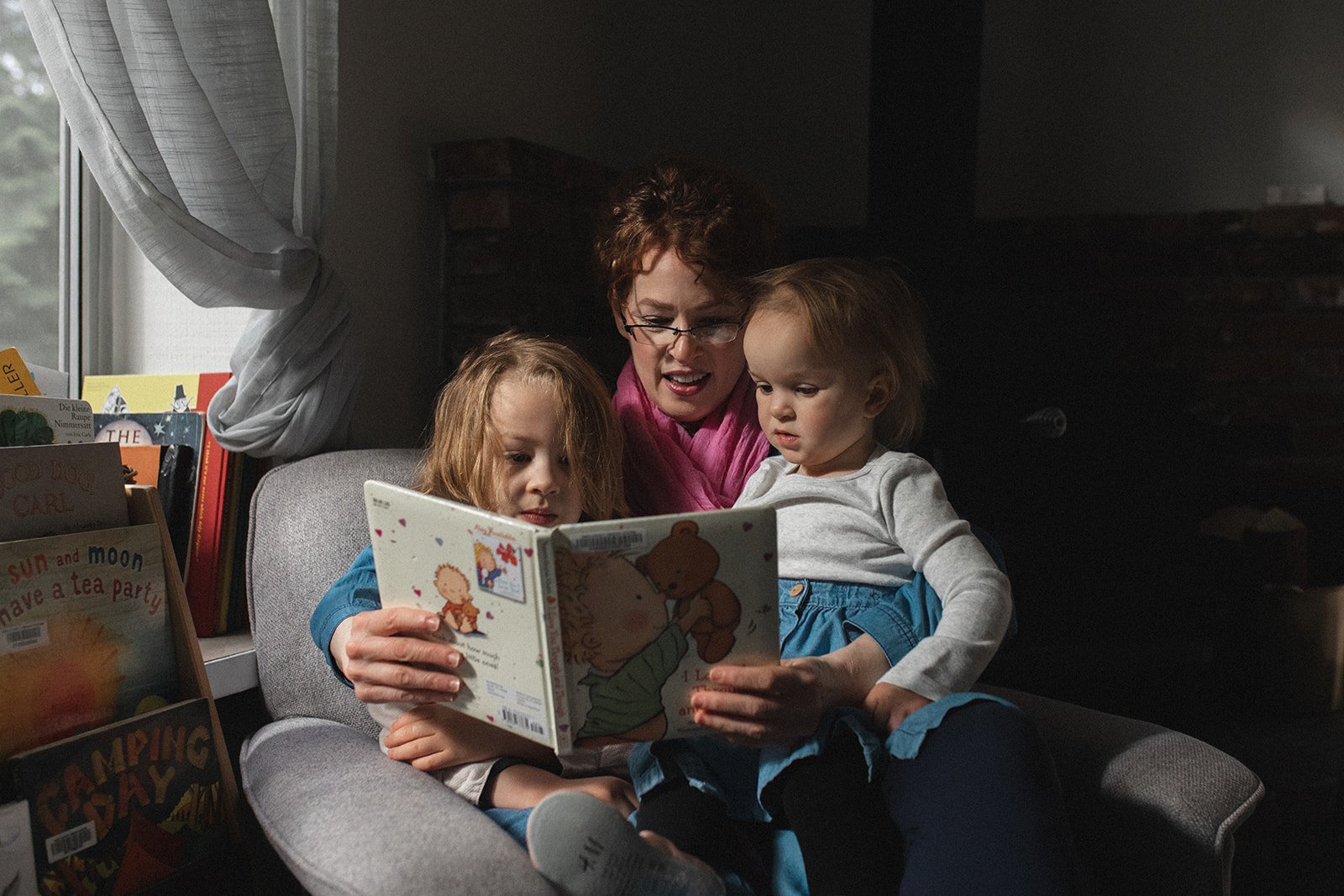 woman with glasses holds two children on her lap while she reads to them