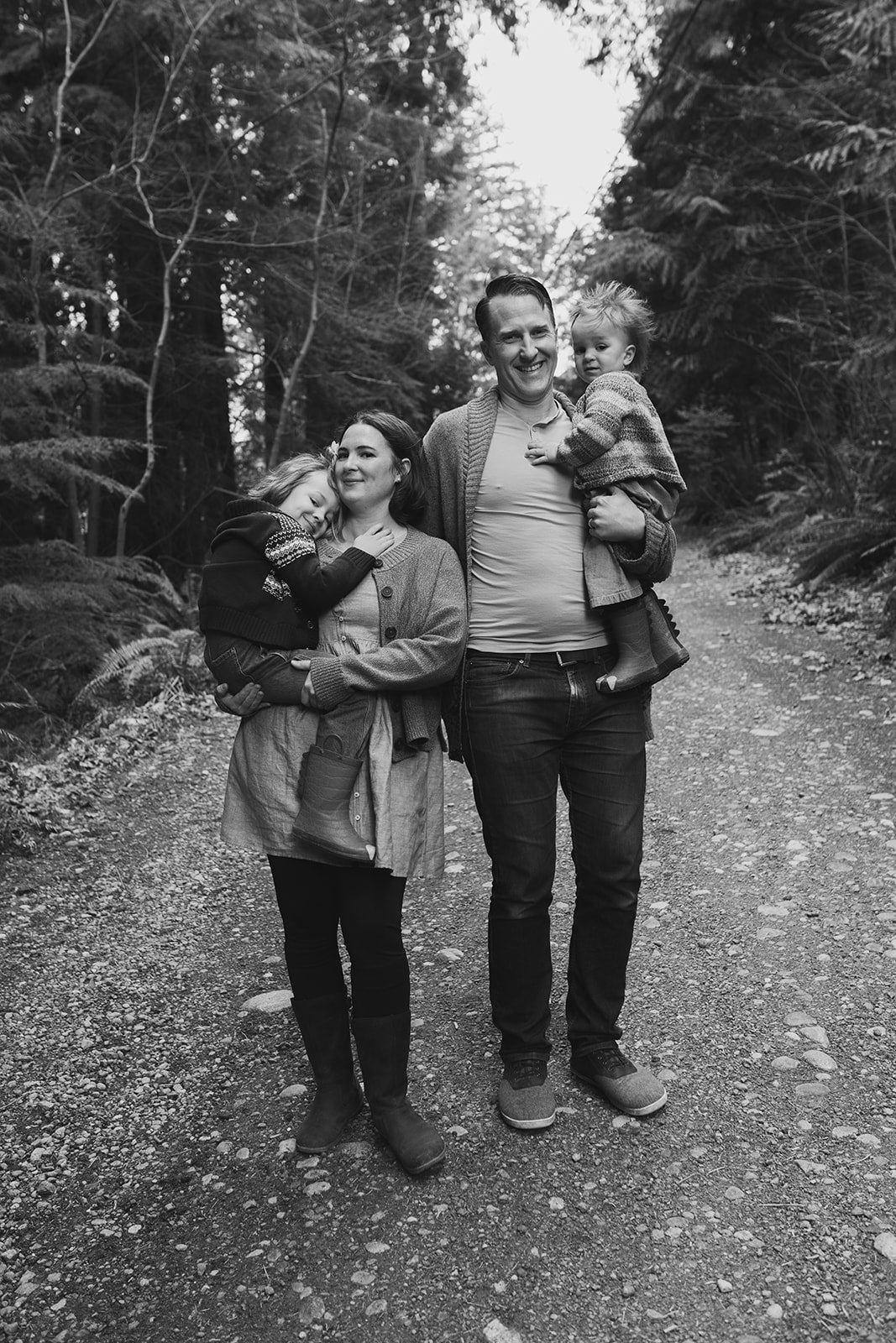 family portrait, mother and father hold one child each in their arms while standing on a path in the woods