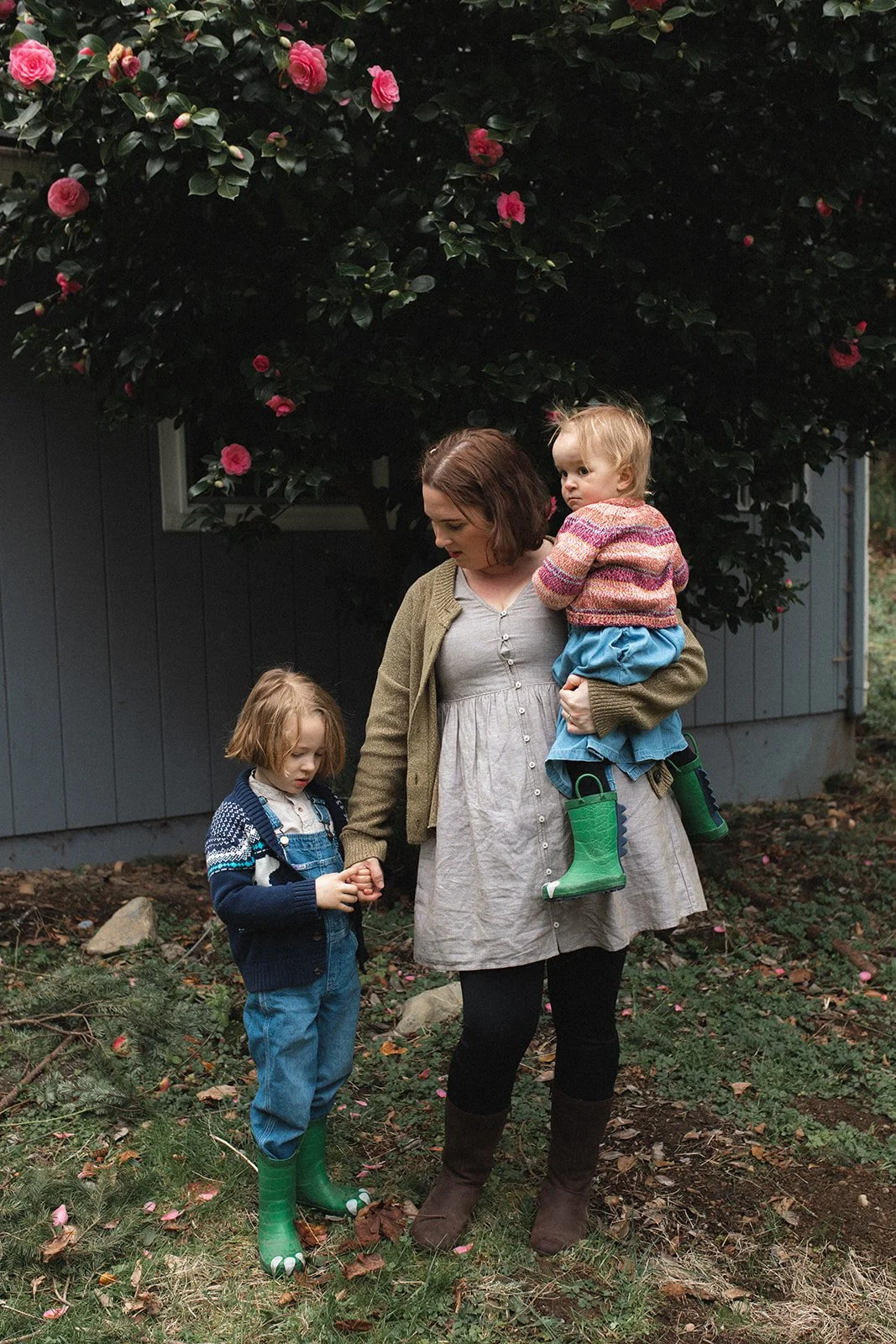 mother stands in grass holding little boys hand and holding little girl in arms