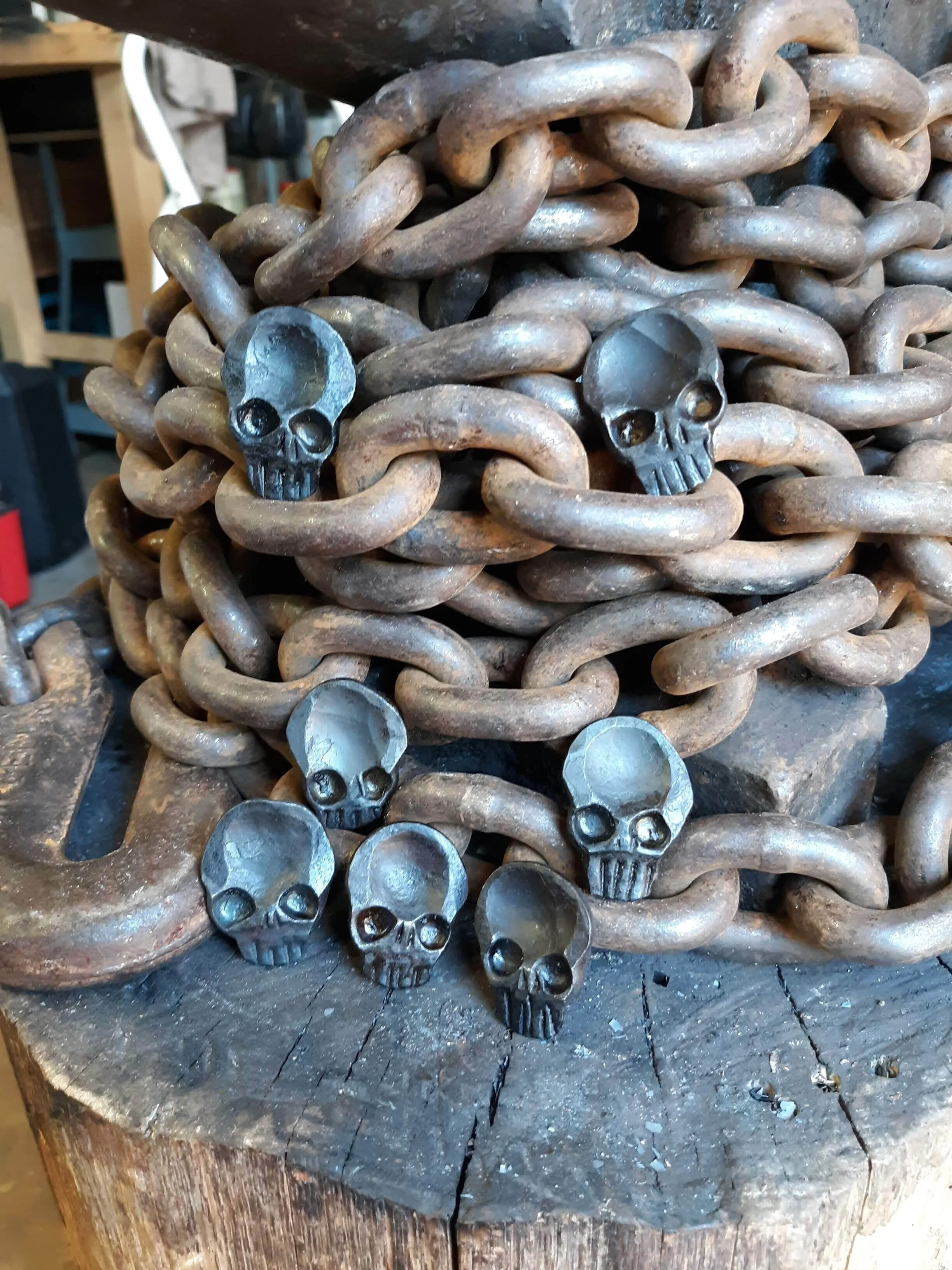 A "Memento Mori" worry coin, hand forged from 3/4" round stock steel and shaped in the form of a skull. Shown here as many coins resting on the chains of the anvil.