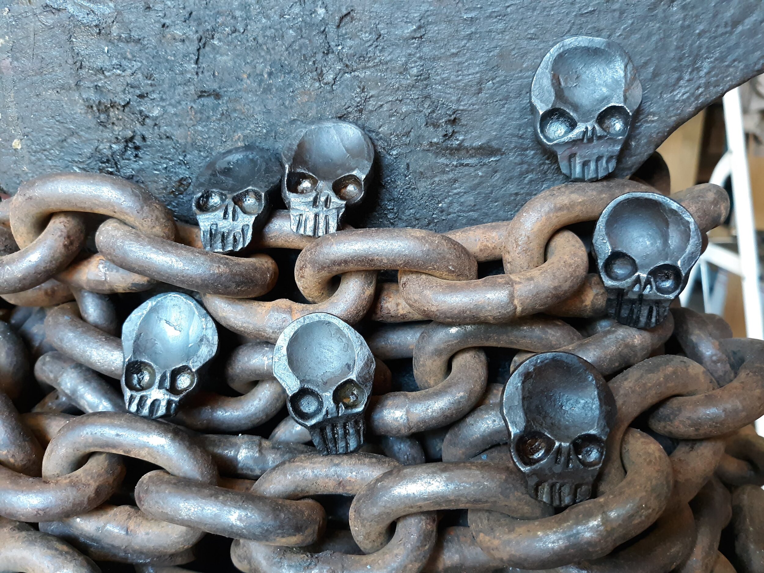 A "Memento Mori" worry coin, hand forged from 3/4" round stock steel and shaped in the form of a skull. Shown here as many coins resting on the chains of the anvil.